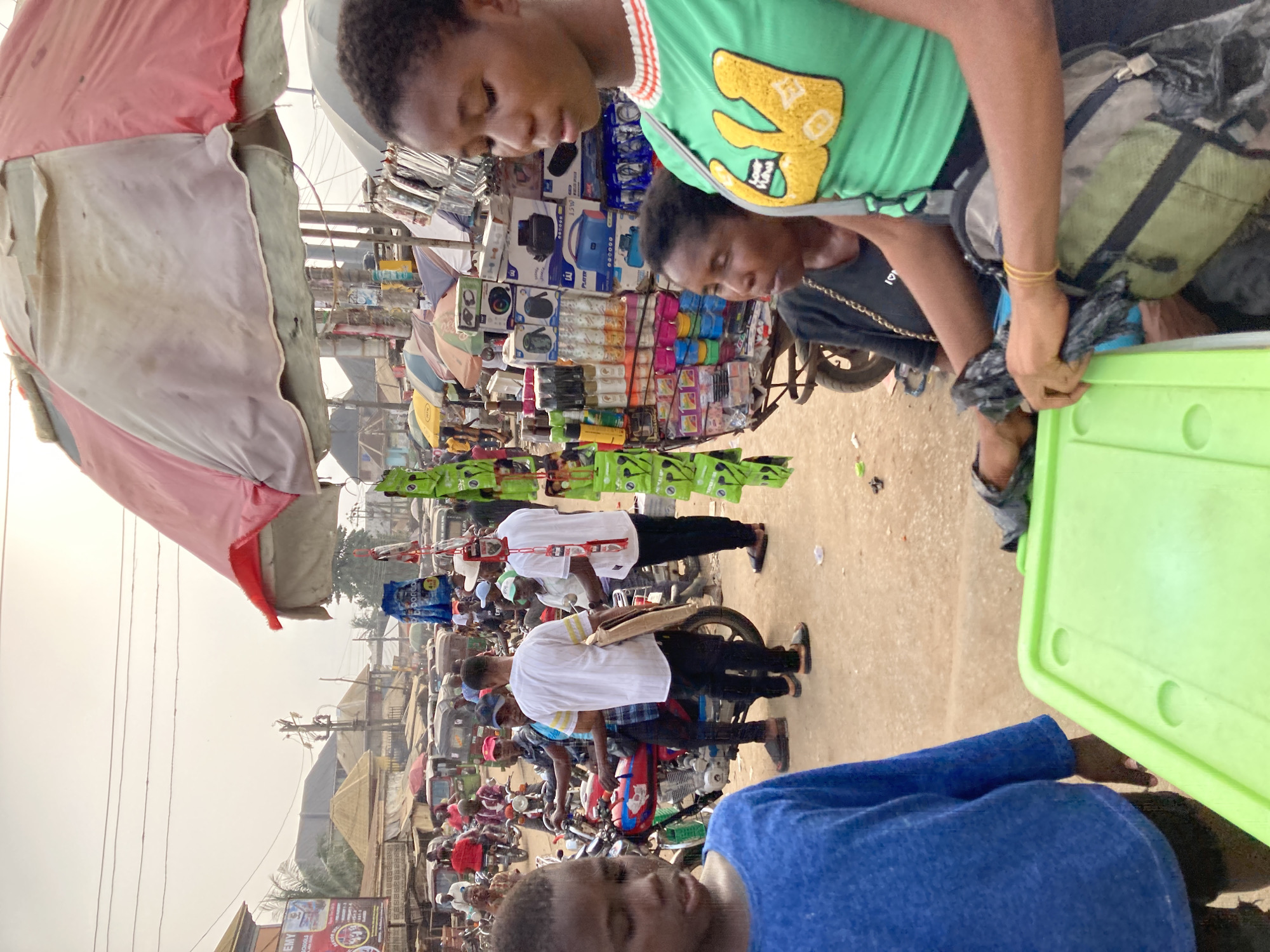 The image shows a street scene in Ura Nta Umuarandu, Nigeria. Two young people, one in a green shirt and the other in a black shirt, are standing next to a cart displaying items for sale. A person in a blue shirt is visible in the foreground. In the background, several people are on motorcycles. There is a pink and white umbrella over what appears to be a market stall. The street appears crowded with people and motorized vehicles.