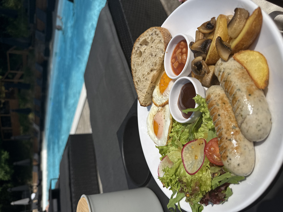 An overhead-angled view shows a breakfast plate positioned poolside in Hatne, Ukraine. The white oval plate features two grilled light-colored sausages, two fried eggs with intact yolks, and a slice of multigrain bread. Accompanying these are a side salad composed of mixed greens, sliced radishes, and a cherry tomato, along with roasted potato wedges and sautéed mushrooms. Two small white ramekins on the plate contain dark brown sauce and baked beans. The plate rests on a dark-colored outdoor lounge chair or low table with a dark grey cushion. A light-colored container with a wooden lid is visible to the left. In the background, a large, blue swimming pool is visible, with dark lounge chairs and light cushions arranged around its perimeter. Several individuals are partially visible in the pool. Beyond the pool, green foliage and distant wooden structures or cabanas are present under bright sunlight.