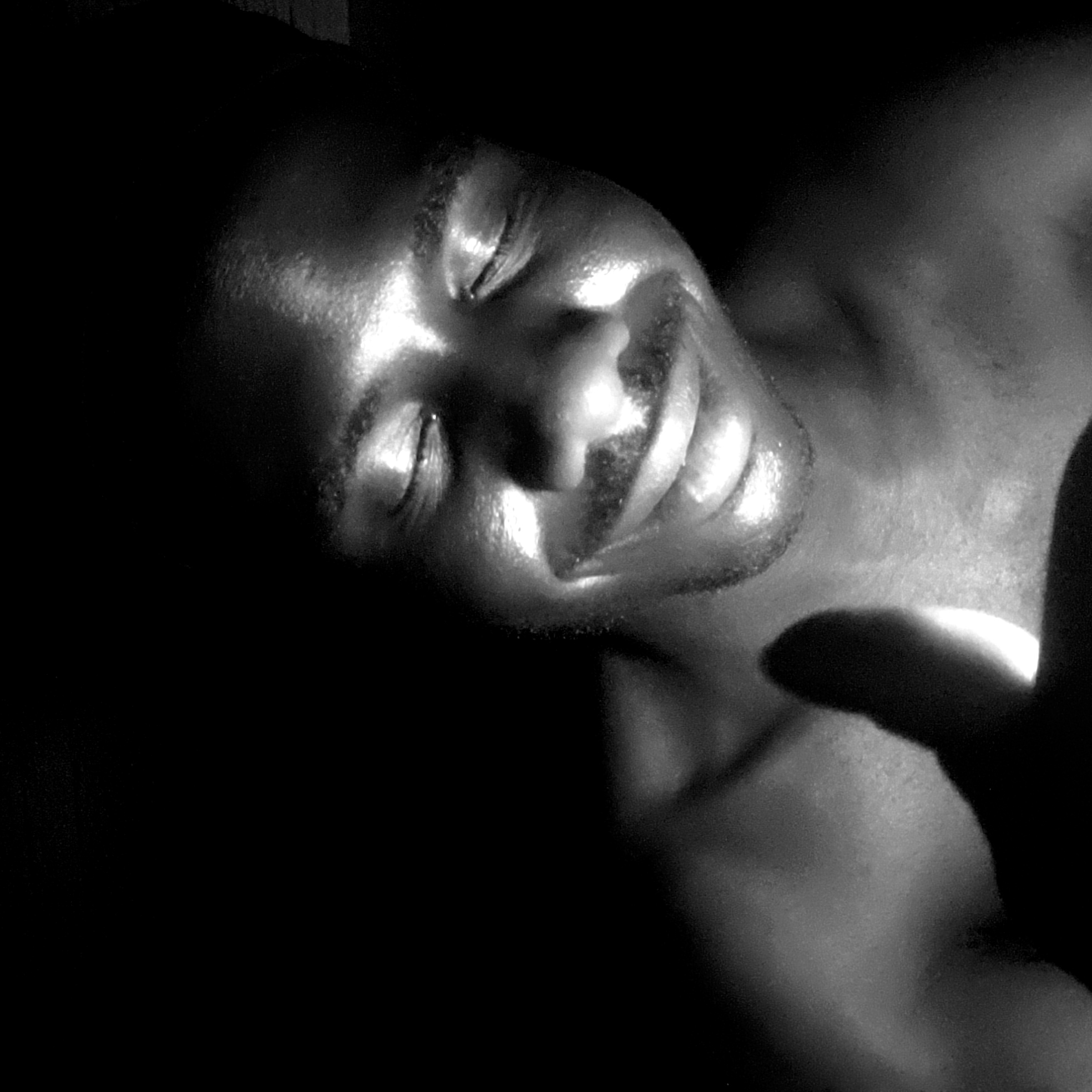 A close-up, grayscale image shows a man lying down. His eyes are closed and he has a slight smile. The light source appears to be directly above and in front of him, creating significant highlights on his face and chest. Part of what seems to be a hand is visible in the lower right corner, casting a shadow. The background is dark. There is no visible indication to place the image specifically in Narayi, Nigeria.