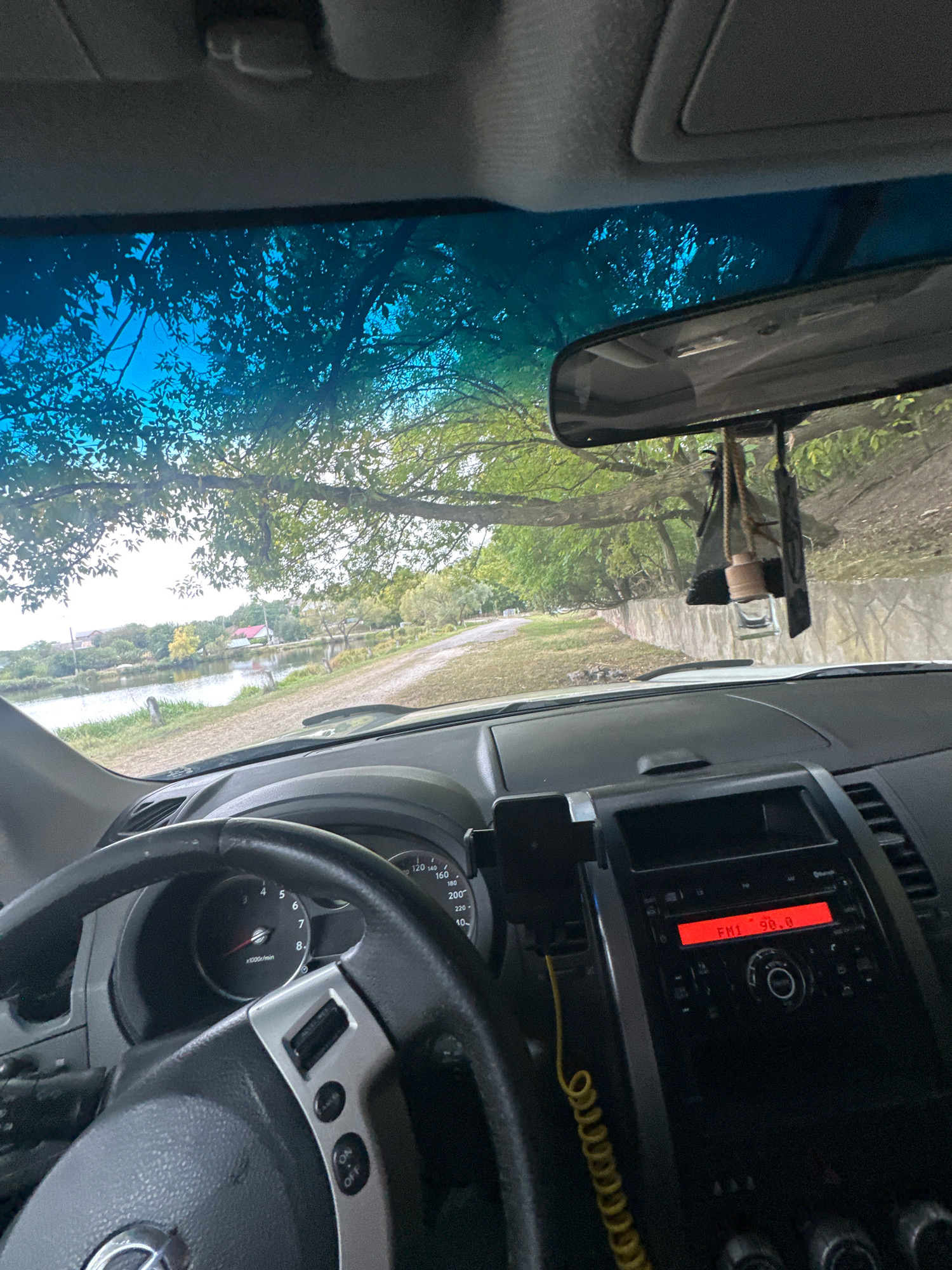 The image is a first-person perspective from inside a car, looking forward through the windshield. The interior of the car is visible, including the steering wheel, dashboard with instruments, and the center console with a car radio displaying "FM1 90.0". A car air freshener hangs from the rearview mirror. Outside, a dirt road leads towards a body of water with trees and houses on the opposite bank. The scene appears to be in Makiv, Ukraine, given the context provided.