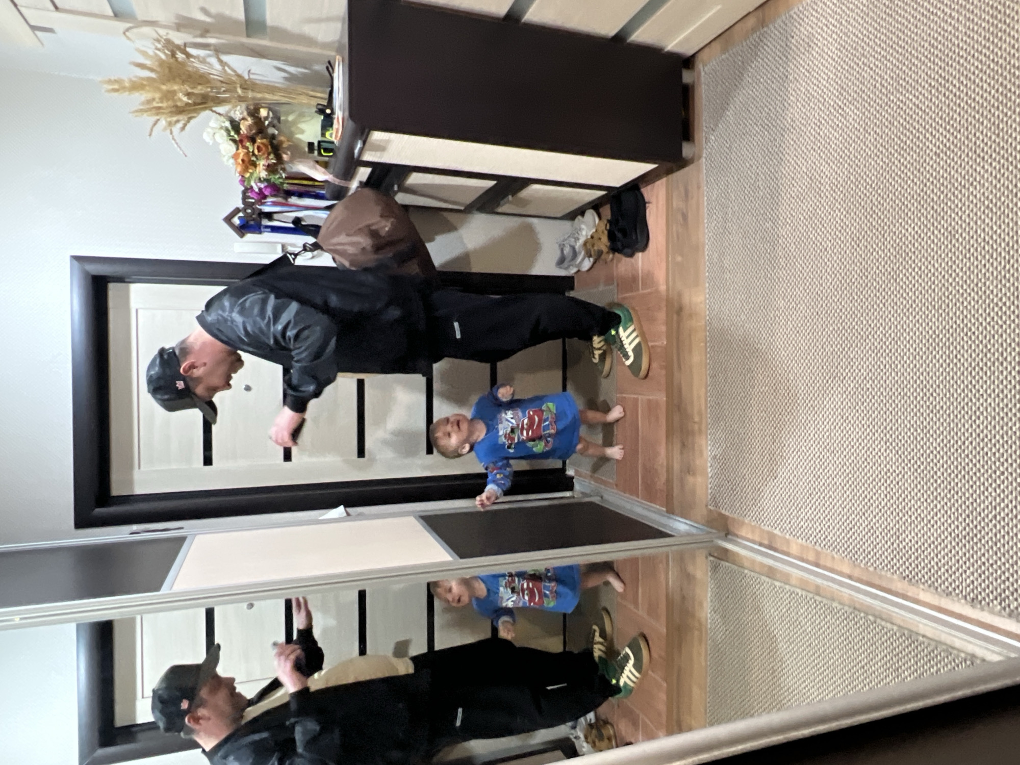This image captures an indoor scene in a hallway or entryway in Noviy, Russia, featuring a man and a young child standing before a large mirrored wardrobe.
The man, positioned on the left, is wearing a dark, long-sleeved jacket, black pants, and a dark baseball cap. He is looking downwards and slightly to his right, seemingly interacting with the child. He wears light-colored sneakers with green stripes. Beside him, a young child stands barefoot, dressed in a blue long-sleeved t-shirt featuring cartoon cars, notably characters resembling Lightning McQueen and Mater from "Cars." The child is looking up at the man, with an expression that suggests engagement or speech.
Behind the man and child, a dark wooden door with light-colored panels is visible. To the right of the door, a dark brown console table or cabinet is placed against the wall. On its surface, there is a decorative arrangement of dried light-colored reeds or wheat stalks and a vibrant bouquet of artificial flowers in shades of orange, pink, and white. Below the cabinet, on a light brown tiled floor, several pairs of shoes are neatly stored. The tiled floor transitions into a light, textured rug that covers the majority of the floor space on the right side of the image.
The large mirrored wardrobe on the left provides a clear reflection of both the man and the child, effectively showing them twice in the scene. The lighting suggests an indoor setting, likely during the daytime. The visible text consists of the cartoon car characters on the child's shirt. The scene depicts a casual, everyday moment between an adult and a child in a home environment.