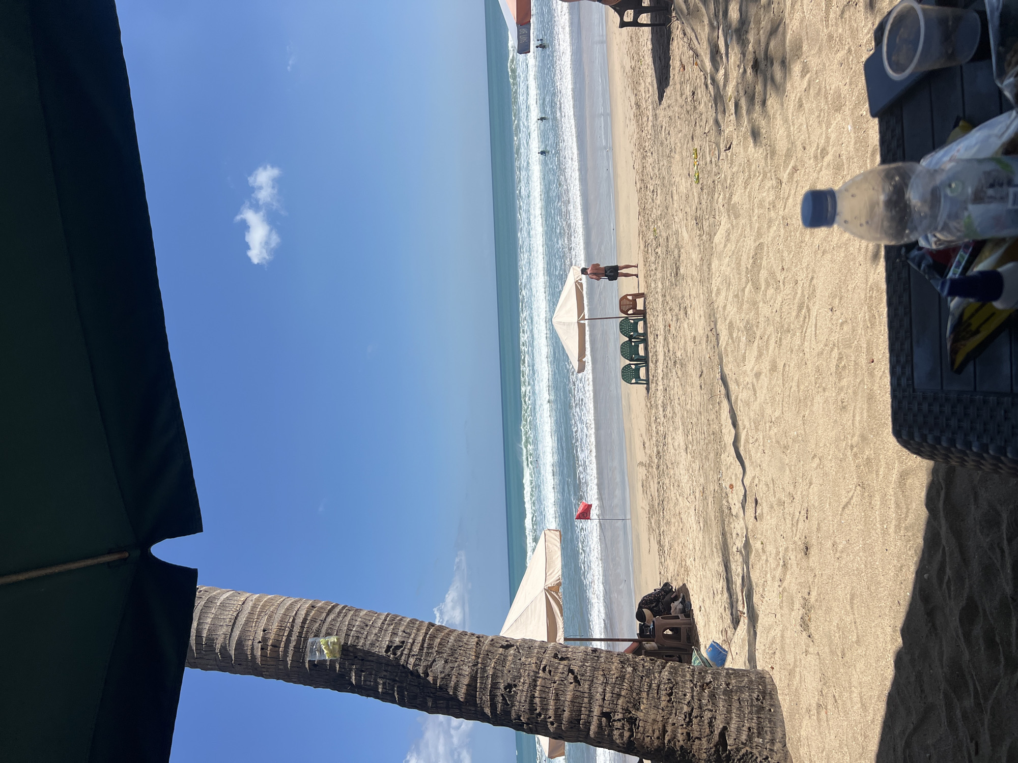 A view of Kuta Beach in Bali, Indonesia, features a sandy shore with the ocean and clear blue sky. In the foreground, the shadow of a dark umbrella covers part of a table with a water bottle, phone, and cup. A large palm tree trunk is on the left. The middle ground shows a few beach chairs and umbrellas, with a red flag planted in the sand. In the distance, people are seen swimming and playing in the waves. The overall scene conveys a typical day at a popular tourist beach.