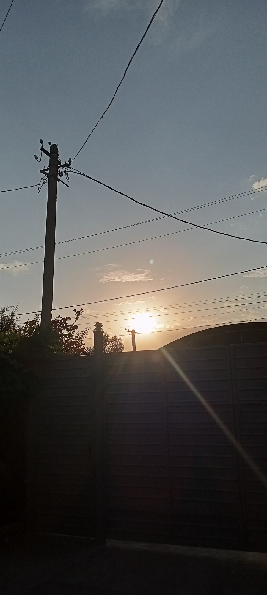 This image captures a sunset scene in Sokuluk, Kyrgyzstan, with a prominent wooden fence dominating the foreground. The fence is dark and horizontally slatted, casting a shadow that covers much of the ground in front of it. Above the fence, the sky is a gradient of blues and oranges, indicating the setting sun, which is partially obscured by clouds and a distant electrical pole. Power lines crisscross the sky, some appearing taut and others slightly saggy. To the left of the frame, some dark foliage, possibly trees or bushes, can be seen peeking over the fence. The overall atmosphere is serene, with the natural light of the sunset creating a warm glow.