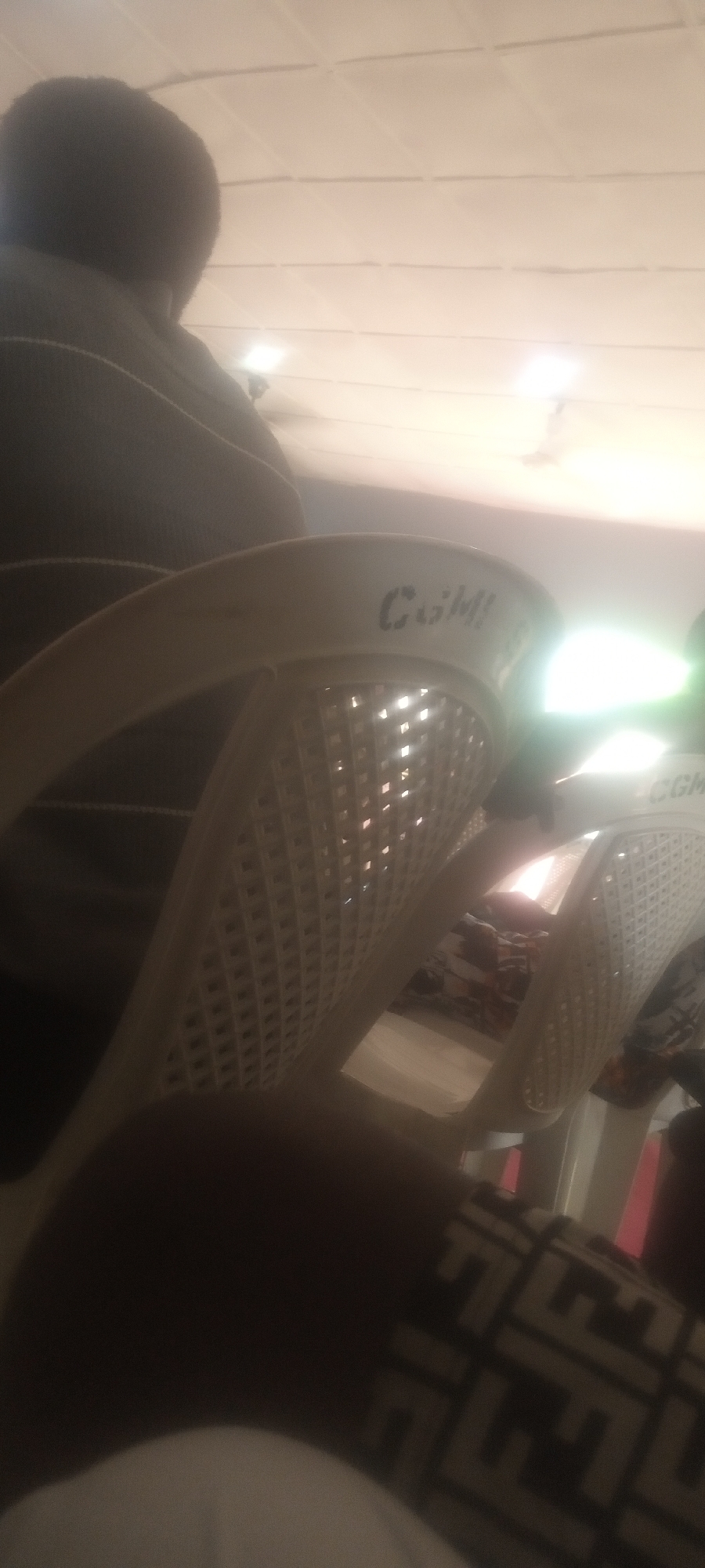 The image shows a person sitting in a plastic chair in what appears to be an indoor space. The chair is white and has the letters "CGM" visible on the upper portion of the backrest. There are rows of similar chairs behind the first one. The person is wearing a dark-colored top and trousers with a complex printed pattern. The lighting in the room is bright. The location is in Sabon Tasha, Nigeria.