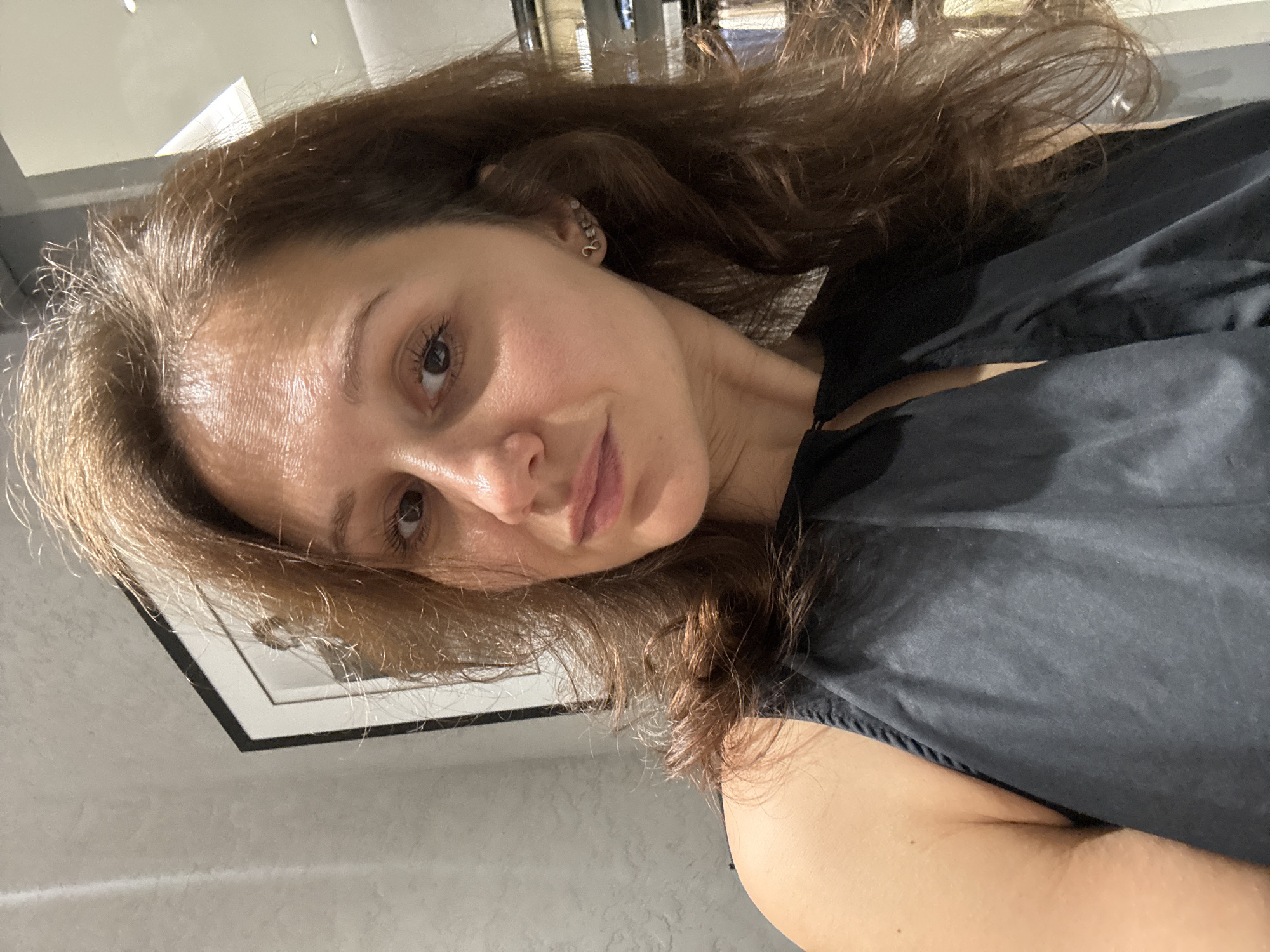 A woman with light brown hair and light skin is wearing a black garment, possibly a salon cape. She is posing for a photograph. She has an earring on her left ear. A picture frame hangs on the wall behind her. The environment appears to be indoors, possibly a salon or a similar establishment. Dubai, United Arab Emirates is not visible in the image.