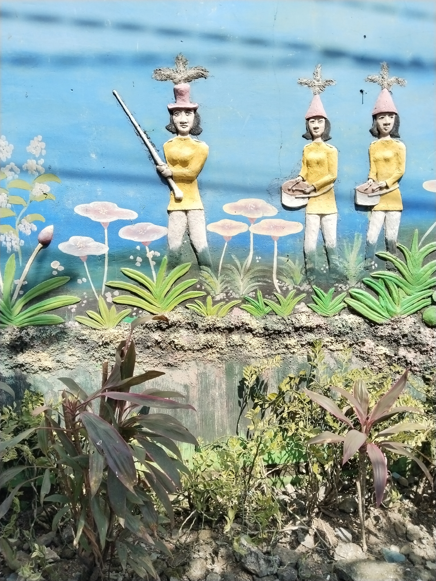 This image depicts a concrete wall with a relief sculpture, likely located in the city of Jirapan, Indonesia. The sculpture features three figures, each depicted in a stylized, somewhat naive manner. The figures are dressed in yellow shirts and white pants, each wearing a hat topped with a decorative element. One figure holds a long object resembling a spear, while the other two appear to be playing drums. The background of the sculpture is painted blue, with stylized white flowers and lily pad-like shapes. The relief itself is painted with bright colors, creating a whimsical scene. The lower portion of the image shows various plants growing in front of the wall, obscuring parts of the sculpture and suggesting an outdoor setting. The wall appears aged, adding to the scene's character.