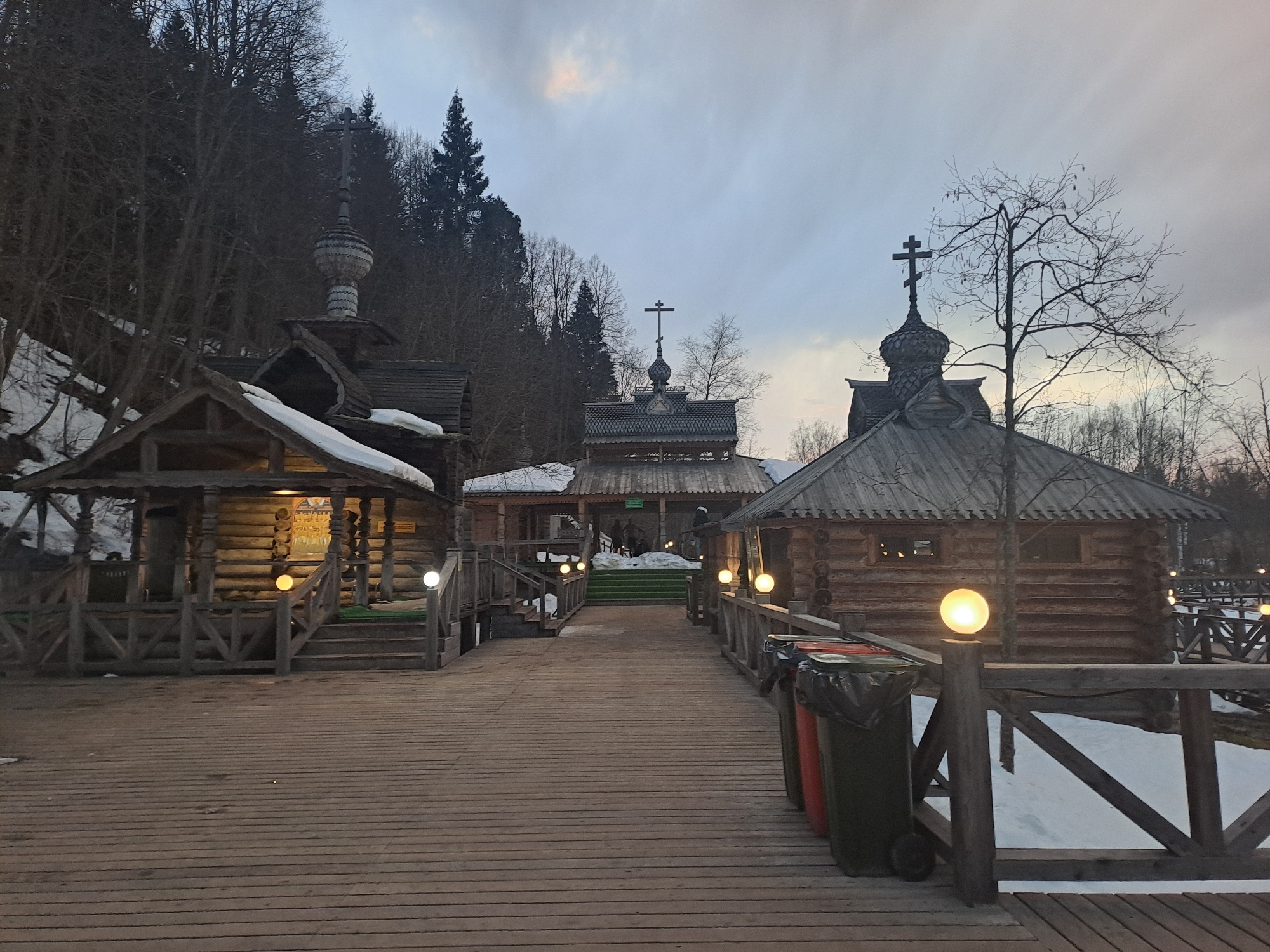 The image depicts a complex of wooden buildings connected by a wooden boardwalk in Vzglyadnevo, Russia. The buildings feature traditional Russian architectural elements, including steeply pitched roofs and crosses atop the structures. Snow is visible on the roofs and in the surrounding area, suggesting a winter setting. The boardwalk is lined with wooden railings and illuminated by spherical light fixtures. Three trash cans are positioned near the boardwalk, adding a mundane element to the otherwise historical scene. Bare trees and a hillside backdrop contribute to the natural environment. The sky is overcast, casting a muted light on the scene. People are visible in one of the buildings in the distance.
