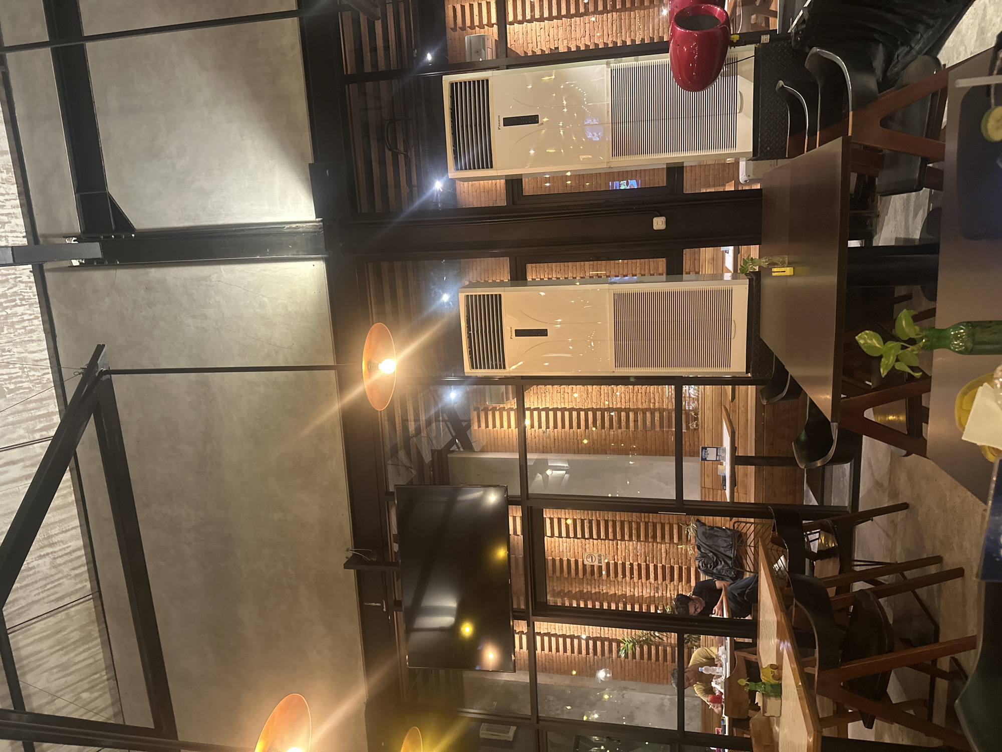 The image depicts the interior of a modern café in Denpasar, Indonesia, possibly within the Seminyak area, judging by the contemporary design. The space features exposed industrial elements like dark metal beams and a concrete-textured ceiling. Large glass windows divide the seating areas from the brick-walled exterior, creating an open and airy atmosphere.
In the foreground, several dark wooden tables with metal legs are arranged, some with chairs. On one table, a small green plant in a glass vase and a yellow object are visible. The lighting is warm, with prominent pendant lights hanging from the ceiling.
Further back, through the glass partitions, more seating areas and individuals are visible. In one section, two men are seated at a table, one appearing to be eating and the other looking down at a book or tablet. Behind them, more tables and chairs are arranged, along with wooden bench seating.
Two tall, white Daikin air conditioning units stand prominently against the brick wall, suggesting a need for climate control in the tropical environment. A large, dark flat-screen television is mounted high on the ceiling beam. A striking red, jet-engine-like object is visible on the right side of the image, its purpose unclear. The overall ambiance suggests a relaxed and contemporary dining or social space.