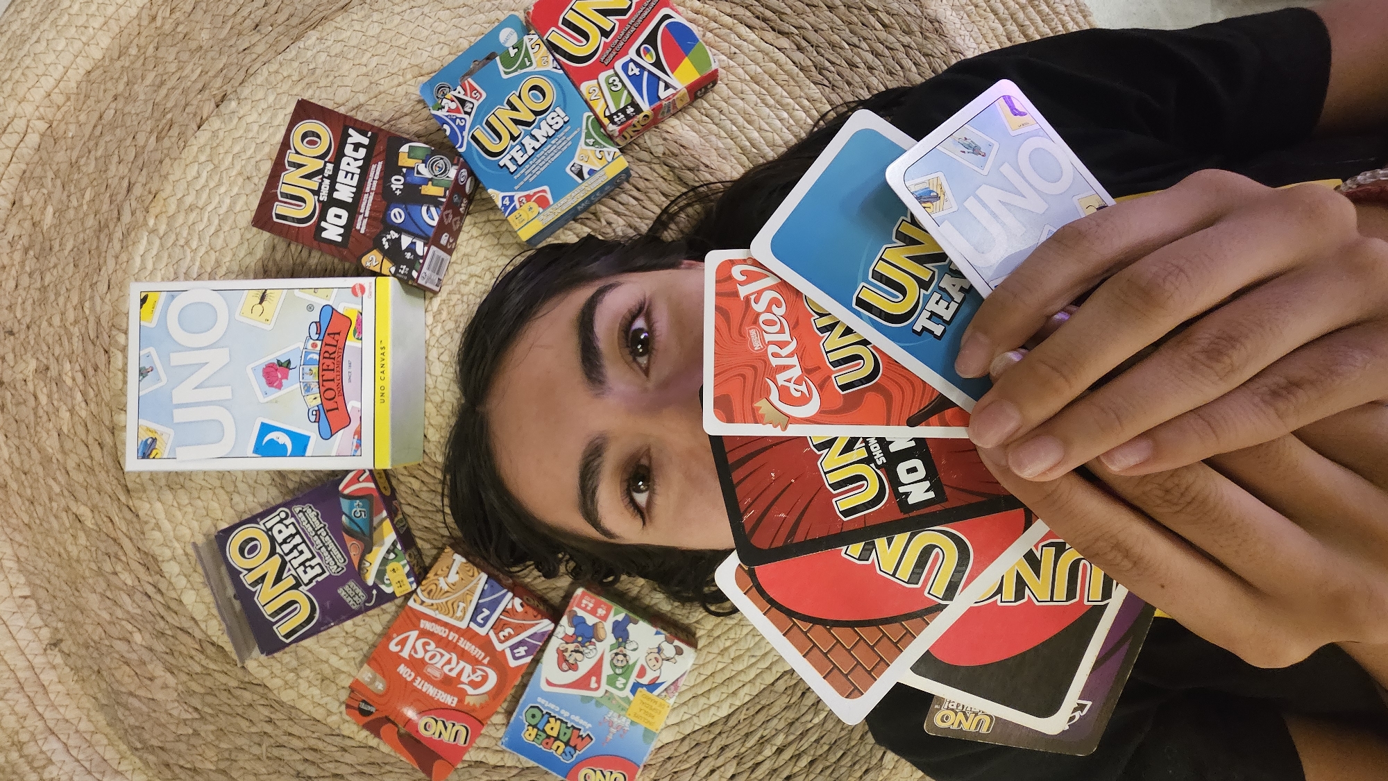 A person lies on a woven mat surrounded by Uno card game variants. The person's head is centered in the frame with their eyes open and looking at the camera. Their hands are holding a stack of Uno cards close to their face. A variety of Uno card game boxes are scattered around the person's head. The games include "Uno Teams," "Uno No Mercy," "Uno Loteria," "Uno Flip," "Uno Carlos V," and "Super Mario Uno." A small part of a black shirt is visible on the right side of the frame. The mat appears to be made of woven natural fiber. The brands "Nestle" and "Don Clemente" appear. There is nothing that can be used to associate this image to the city of Puebla.