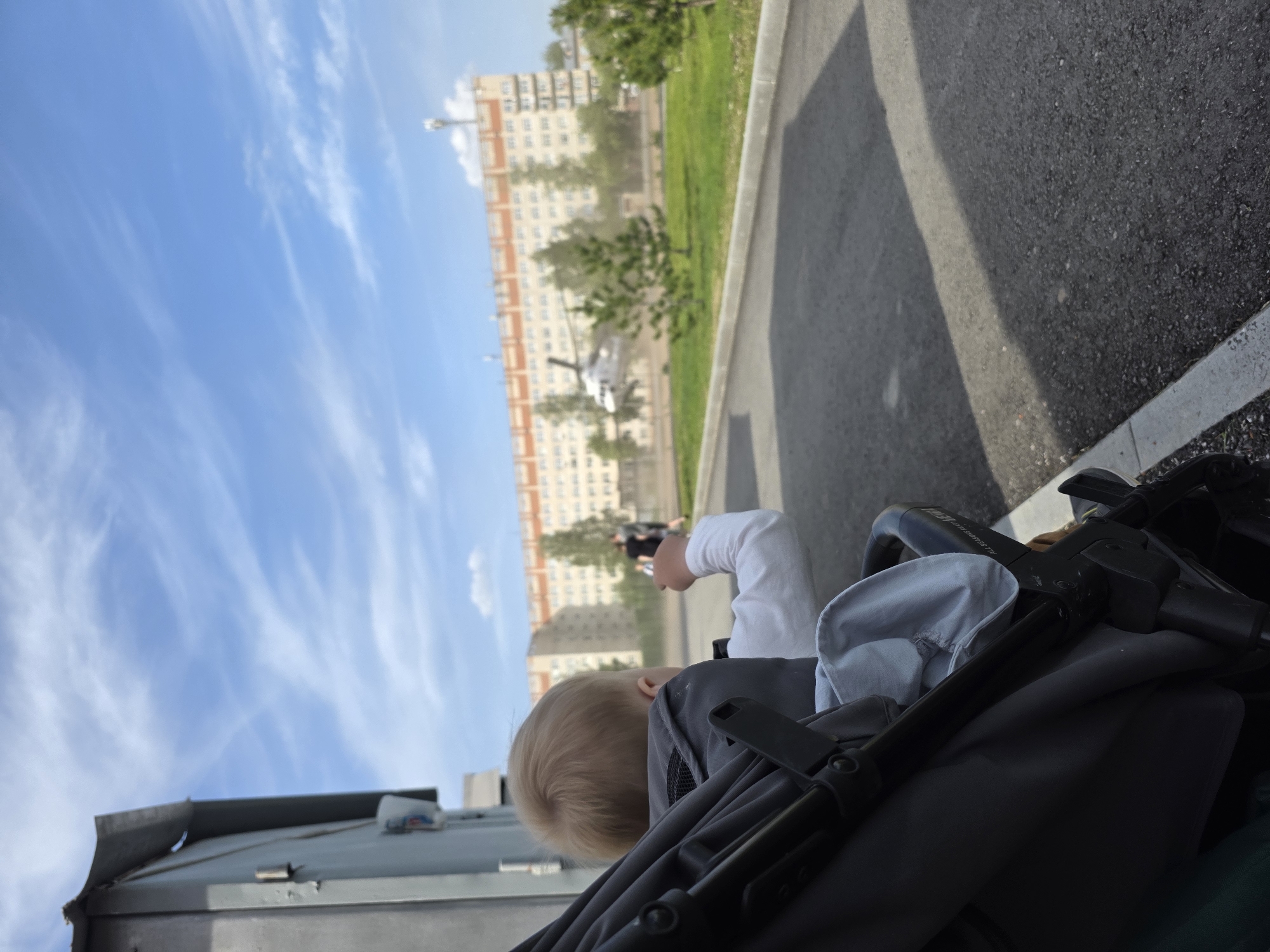 A photograph captured from a low angle shows a child in a stroller, with a cityscape in the background under a sky with light, streaky clouds. The stroller, primarily black and gray, dominates the foreground, with only the child's blond hair and part of their white-sleeved arm visible. In the midground, the cityscape features a tall, light-colored building with many windows, a patch of green grass, and a helicopter. A couple of figures can be seen walking in the distance. The ground near the stroller is asphalt. The location is Unknown, Unknown.