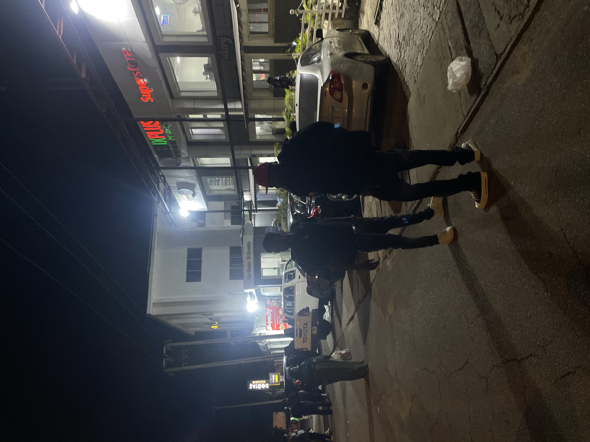 The image depicts a street scene in Abuja, Nigeria at night. The location is identifiable by the "Superstore" and "IXPLUS" signage on a multi-story building. Two individuals are prominent in the foreground. One is wearing a red cap, dark jacket, and black pants, while the other is wearing dark clothing. Both appear to be standing on the sidewalk. Parked vehicles, including a white car and a Toyota pickup truck, are visible along the street. In the distance, there are additional people and storefronts with illuminated signs. The ground shows paved surfaces with cracks and wear. A discarded plastic bag rests near the curb.