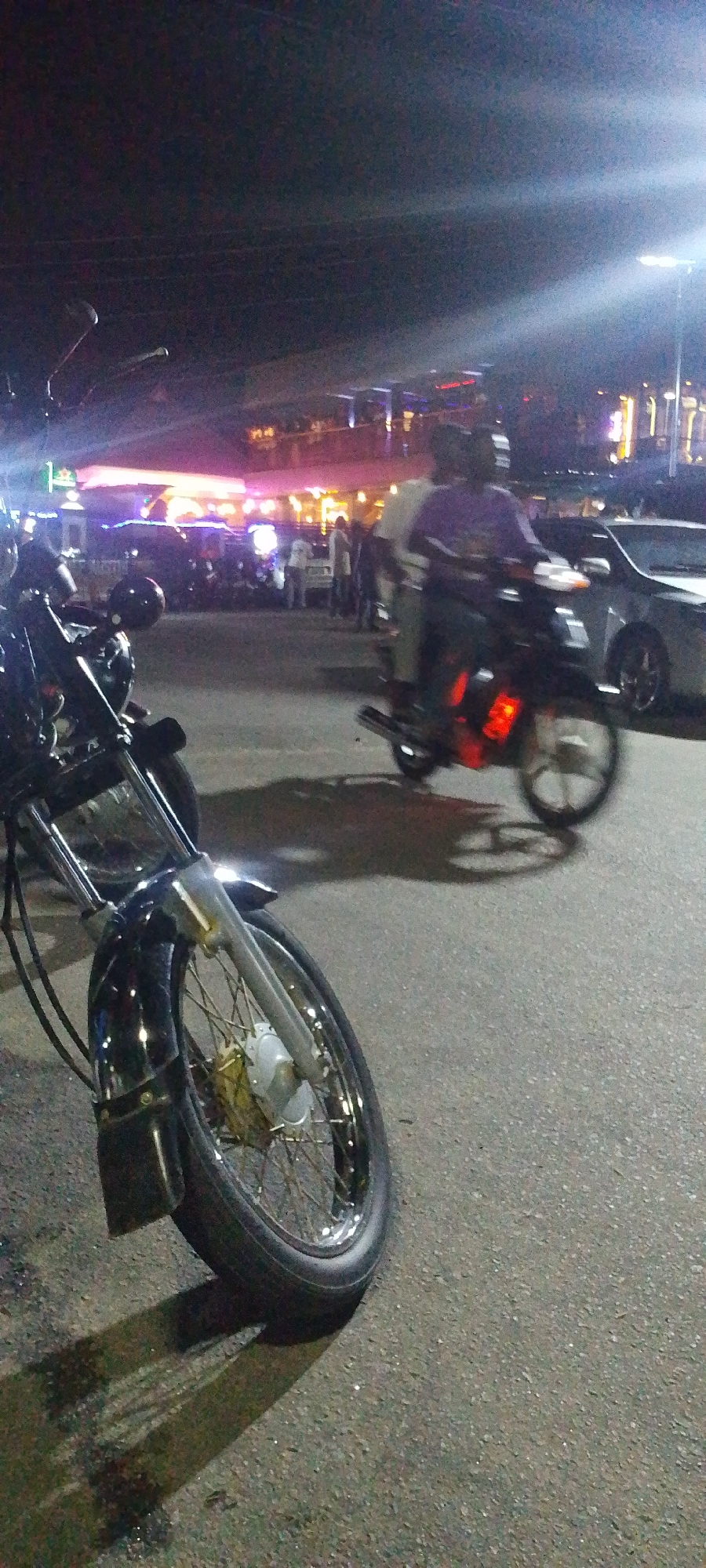 The image shows a street scene at night in Lokoja, Nigeria. In the foreground, there is a motorcycle parked on the side of the road. The front wheel is visible. In the background, there are people gathered near a building with bright lights. A second motorcycle with two people on it is passing by, blurring due to motion. There are cars parked nearby as well. The environment appears to be a commercial or entertainment area.