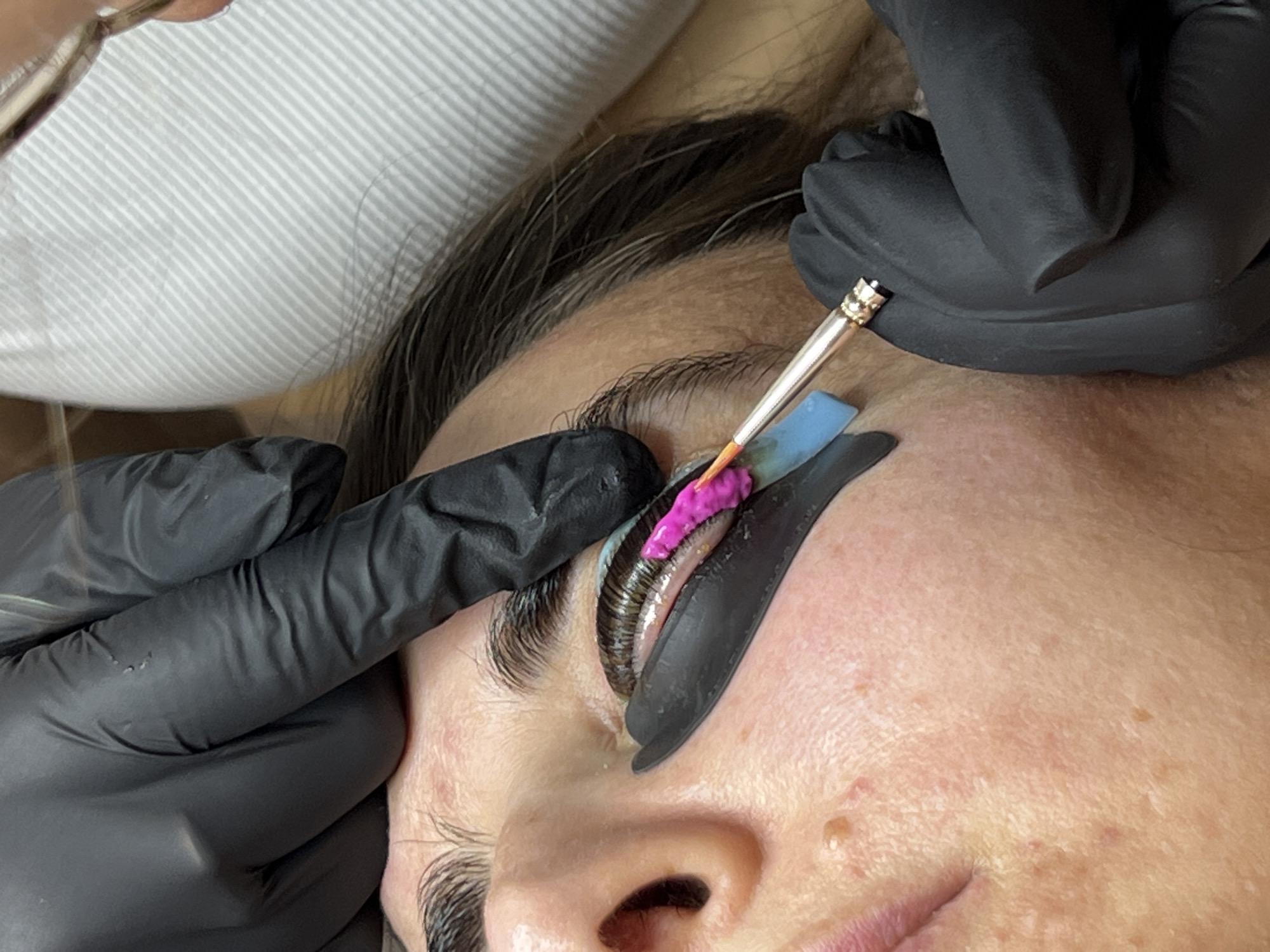 A close-up shows a person's face undergoing a lash lift treatment. The person's eye is closed with a black silicone shield positioned above the upper lash line. The eyelashes are adhered to the shield and coated with a pink-colored product being applied with a thin brush held by a hand wearing a black glove. Another hand wearing a black glove stabilizes the eyelid. A blue strip is positioned between the eyelid and the lash shield. The person has fair skin with freckles and is lying on a white striped surface. The location is possibly in a beauty salon or a private residence in Podgorica, Montenegro, but there is no evidence to confirm this.