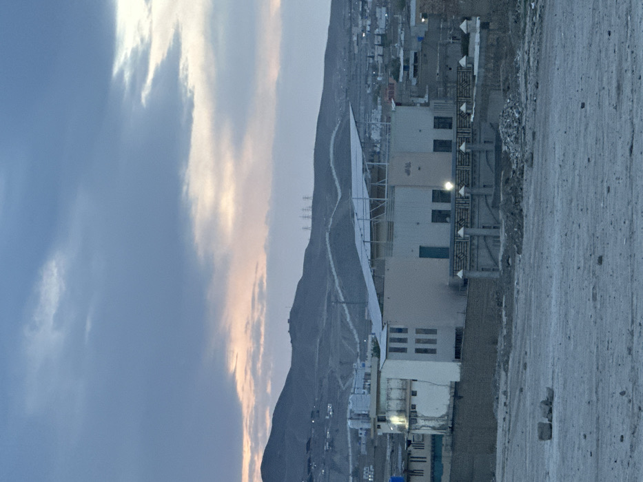 The image depicts a semi-urban landscape in Quetta City Tehsil, Pakistan, observed during dusk or dawn under an overcast sky with subtle pink and orange light near the horizon. The immediate foreground consists of barren, light-colored earth with scattered debris. In the midground, a prominent two-story or taller building, likely a mosque or community center, features a light-colored facade. Its roof is equipped with a large array of solar panels supported by a metal framework. The building displays multiple rectangular windows, a turquoise-colored door, and Arabic script, possibly "Allah," on an upper exterior wall, which is illuminated by a small light source. A decorative, pillared fence with two illuminated lights borders the building's immediate perimeter, behind which a pile of rubble is present. To the left of this main structure, a weathered, light-colored building with multiple vertical window openings is visible. Beyond these structures, a dense arrangement of low-rise buildings characteristic of an urban settlement is observed, including one with a blue tarp on its roof. The background is dominated by a range of dark, undulating hills and mountains. A distinct light-colored, winding structure, possibly a wall or terracing, traverses the hillsides. Multiple electricity transmission pylons are visible on the distant mountain ridges. Scattered artificial lights are discernible across the hills, indicating distant habitation. No people are visible within the frame, and no specific actions or interactions are depicted.