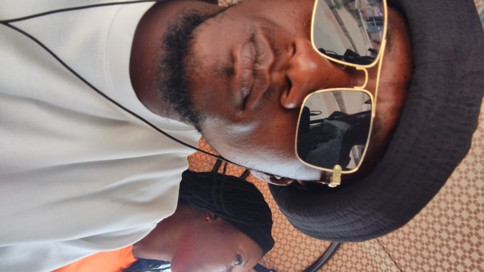A medium shot depicts a person with dark skin and hair, wearing sunglasses and a dark hat, lying down. They are wearing a white shirt. Another person, possibly female, with dark braided hair is partially visible at the bottom of the frame. The surface beneath the person has a patterned design. The reflection in the sunglasses appears to show an indoor setting. There's no definitive information in the image to determine its specific location in Jos, Nigeria.