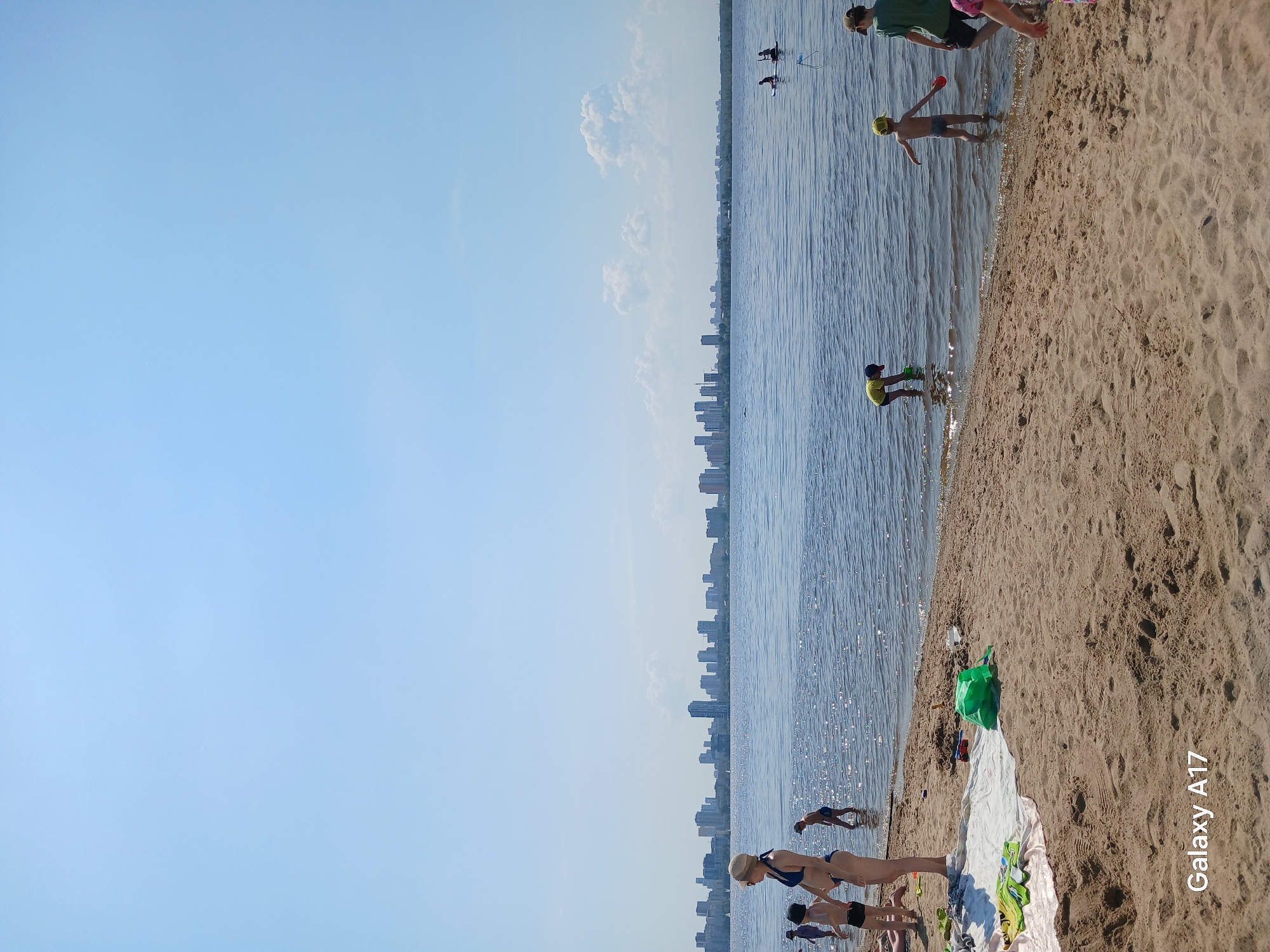 The image shows a beach on the shore of a body of water. Several people are present on the beach and in the water, including children and adults. Some are standing near the shoreline, while others are further out in the water. In the background, a cityscape is visible across the water. The environment includes sand, water, and a clear sky with some clouds. The location is likely in the city of Yekaterinburg, Russia.