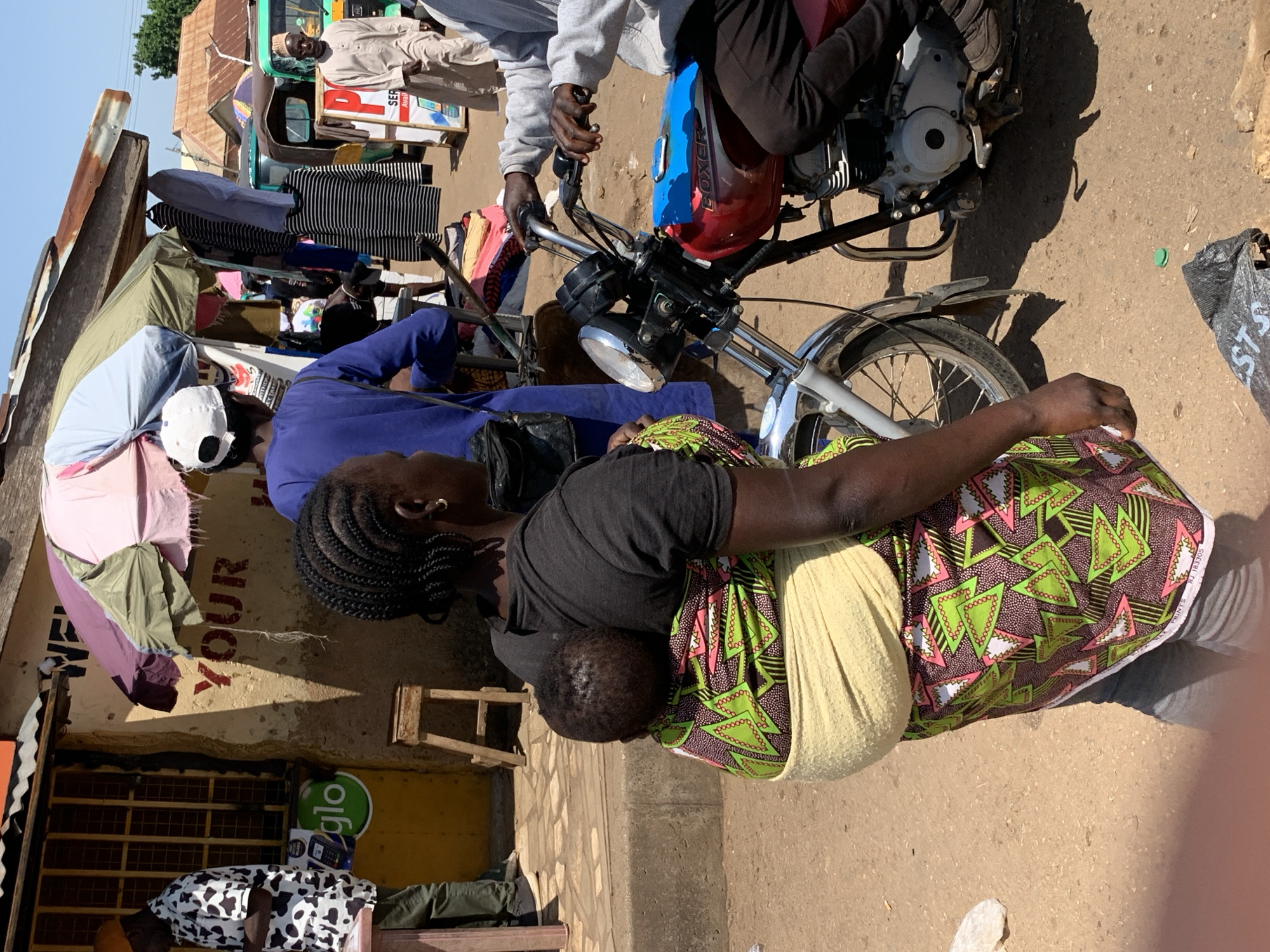 This image captures a bustling street scene in Gura Top, Nigeria, likely a market or commercial area. The foreground is dominated by a woman carrying a baby on her back, secured with a light-colored fabric wrap. She is wearing a dark t-shirt and a vibrantly patterned skirt featuring green triangles with pink accents on a dark background. Her hair is styled in braids, and she has a gold earring in her visible ear. To her right, a motorcycle is partially visible, red with blue accents on the fuel tank, with a rider in a grey hoodie and dark pants. The motorcycle's handlebars, front wheel, and part of the engine are clearly depicted.
In the background, the scene unfolds with more activity. A person in a blue long-sleeved shirt and a white baseball cap stands near a stall with various clothing items hanging. Further back, a man in a light-colored traditional garment walks past a stall displaying cell phones and other goods. A tricycle, commonly used for transport in Nigeria, is parked behind this stall. The environment appears to be a mixture of permanent structures with corrugated iron roofs and temporary stalls with umbrellas providing shade. The ground is dusty and uneven. The overall impression is one of a lively, everyday commerce scene in a Nigerian city.