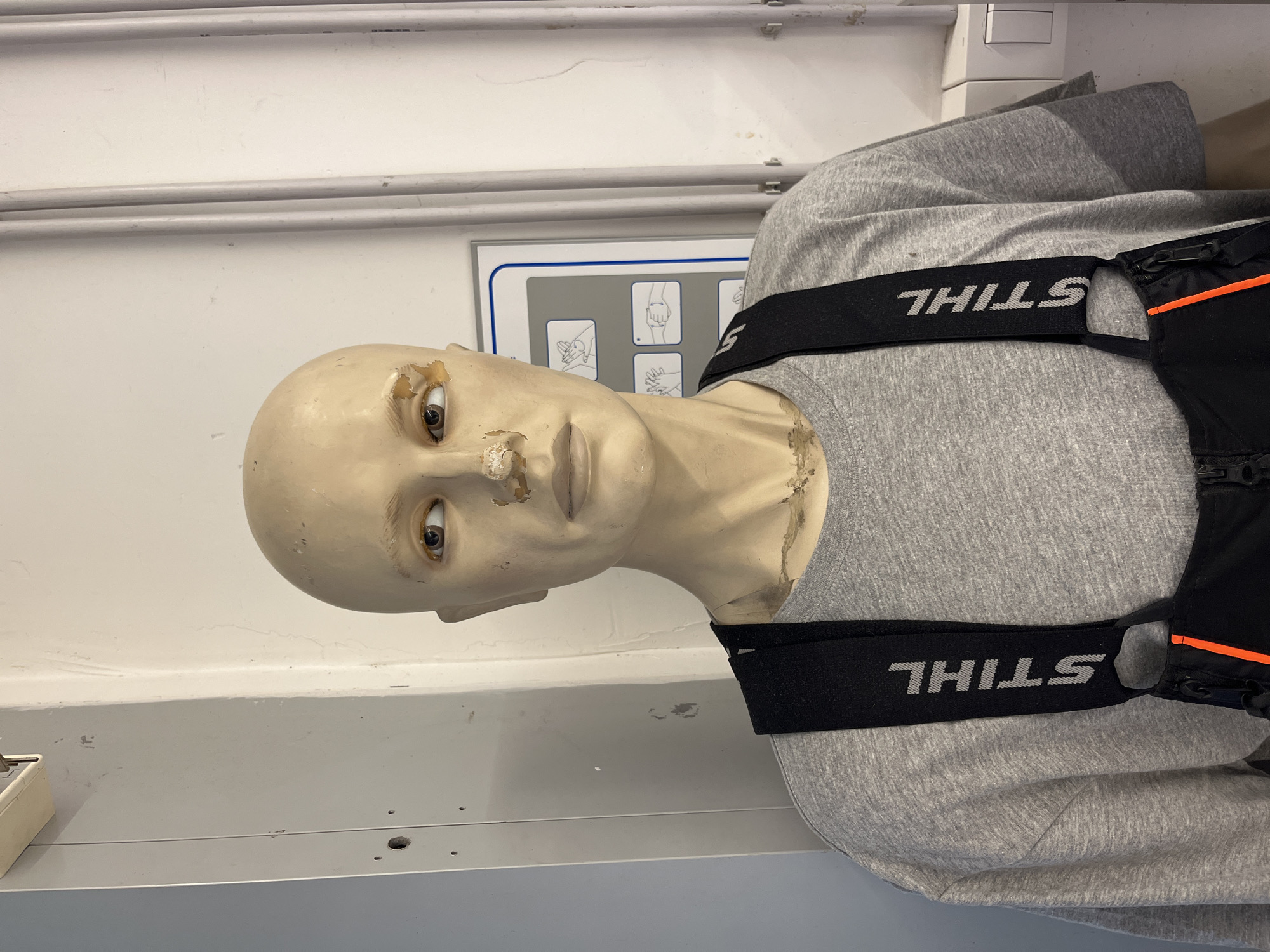 A pale, humanoid mannequin with realistic eyes is positioned in an indoor environment. The mannequin wears a grey long-sleeved shirt and a black vest with "STIHL" printed in white on the straps. The mannequin appears worn, with visible surface imperfections. The background consists of a white wall with a grey horizontal panel and a sign depicting instructions with hand gestures. The location, Budva, Montenegro, cannot be determined from the visual information provided.