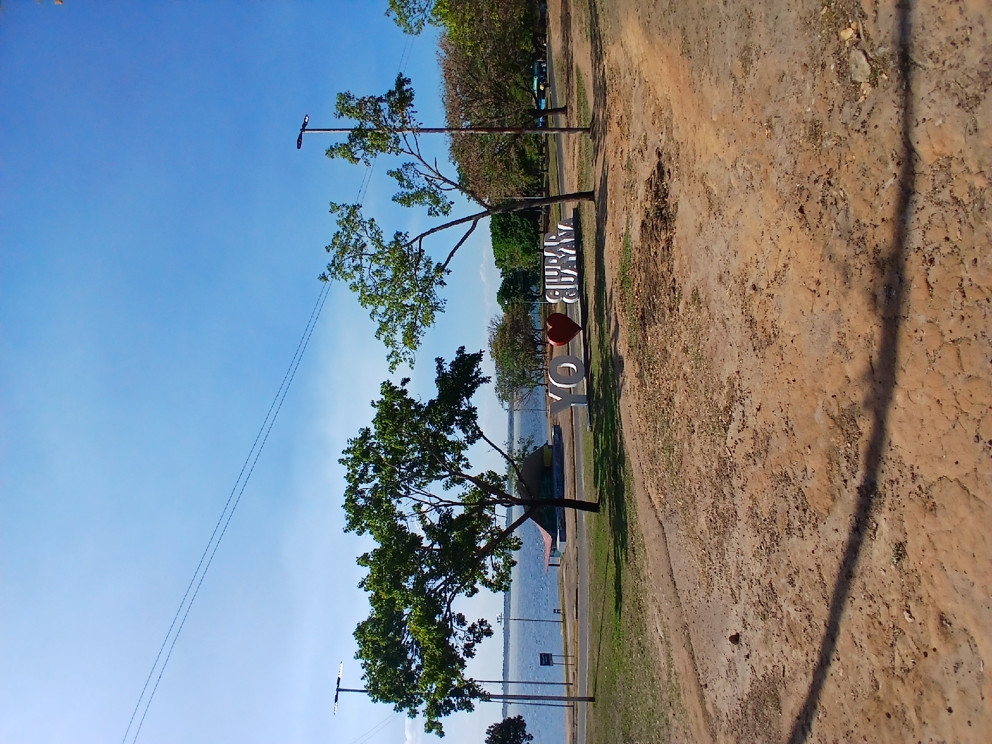A photograph of the "Ciudad Guayana" landmark sign. The sign is white, spelling out the city's name, with a red heart serving as a visual element within the lettering. The sign stands on a grassy area beside a dirt path. In the background, there are trees and a body of water. Above, the sky is clear and blue.