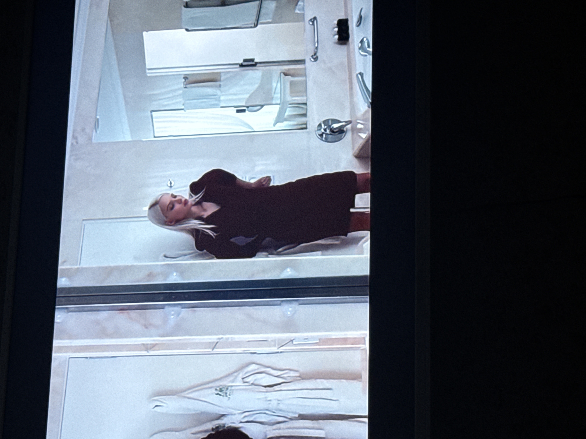 The image depicts the reflection in a mirror of a woman lying on a surface. The woman has blonde hair and is wearing a dark-colored dress. Above her are shelves with towels and bathroom fixtures, including a shower handle. Below her is a shelf with what appears to be a white bathrobe. It's difficult to determine the precise location within the city of Mytishchi, Russia, from this image alone. Additional information or context would be needed to ascertain the specific location.