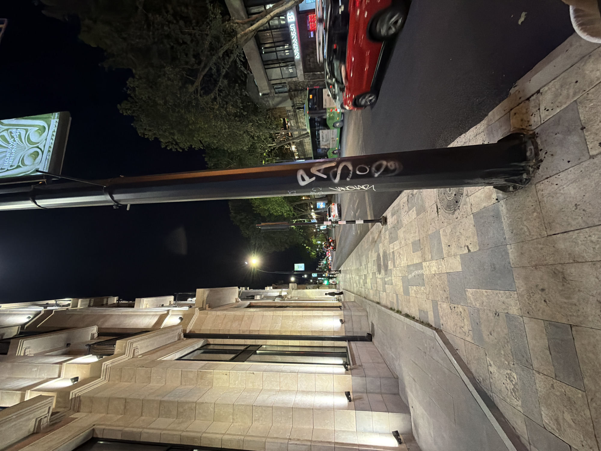 The image depicts an urban scene at night in Tbilisi, Georgia. The view is oriented along Rustaveli Avenue. A streetlight pole, covered in graffiti, dominates the foreground, with a rectangular sign attached near its top. Beyond the pole is a multi-story building with a stepped facade constructed from beige stone blocks. Windows are visible on lower floors.
On the other side of the pole, the roadway and sidewalks are visible. A red car and green bus are traveling along the road. Sidewalks are paved with light-colored stone tiles. Further down the avenue, other streetlights and buildings can be seen in the distance. The sky is dark.