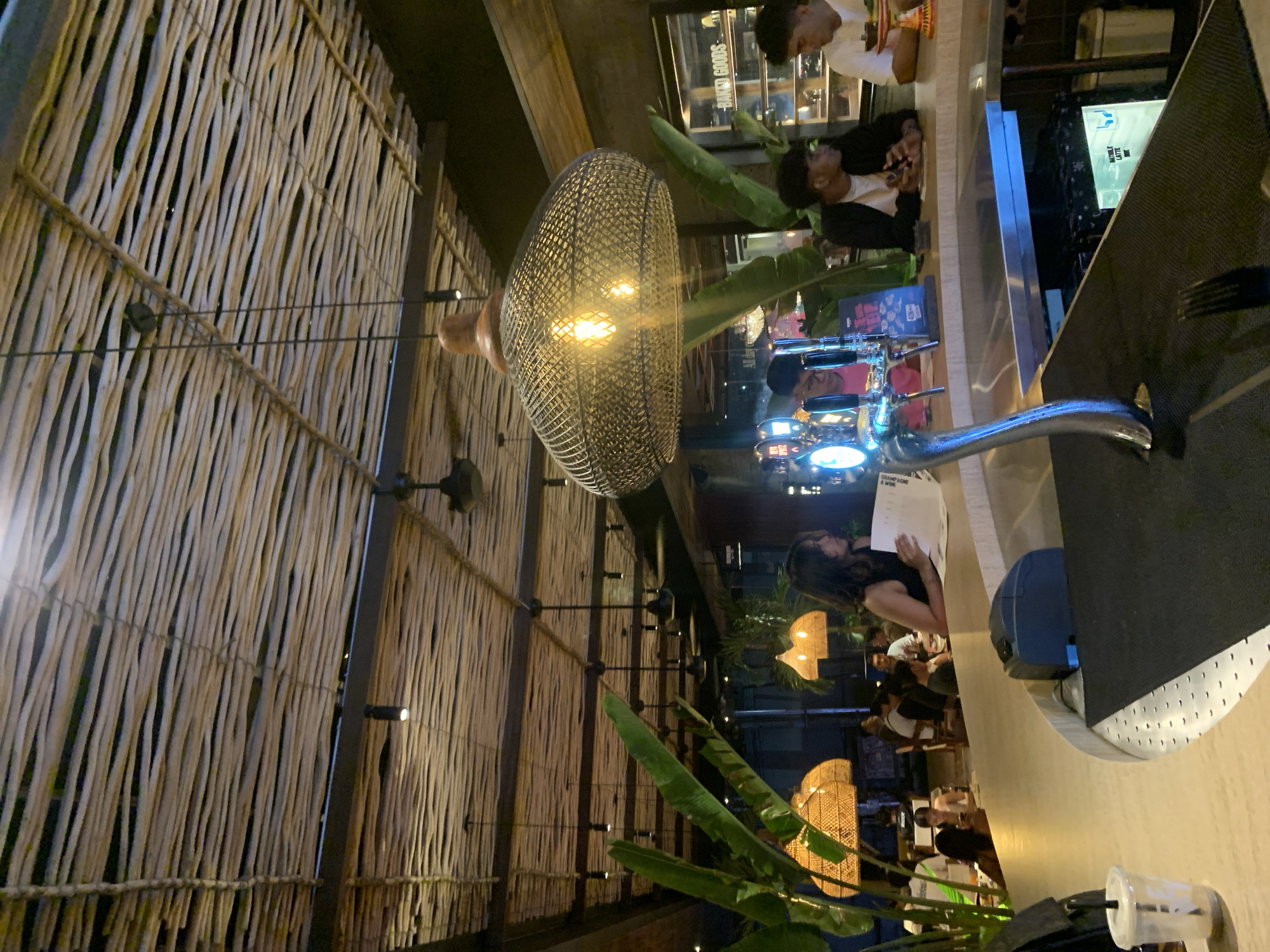 This media file captures an interior scene, likely a restaurant or bar, characterized by warm lighting and natural decor elements, set in North Kuta, Indonesia. The image is oriented sideways.
The setting features a prominent wall or ceiling covered with tightly arranged, light-colored wooden or bamboo sticks, creating a rustic yet modern textured backdrop. Scattered spotlights are integrated into this surface, providing ambient illumination. A large, intricate pendant lamp, crafted from dark metal mesh, hangs centrally, emitting a warm, bright glow from its exposed bulbs. Lush green tropical plants with broad leaves are strategically placed, adding a touch of nature to the environment.
Several individuals are present, seated at a bar or high-top tables. In the foreground, a person with dark hair, dressed in black, is seen holding and possibly reading a menu that displays the text "CHAMPAGNE & WINE". Further back, behind a counter, two young men are visible, one in a white shirt and another in a dark jacket, engaged in conversation or observing their surroundings. Other patrons are partially visible in the background, enjoying the atmosphere, some illuminated by additional, basket-weave style pendant lamps.
The bar area features a sleek, light-colored countertop. A modern silver beer tap system with glowing elements is prominently displayed in the mid-ground. Reflections on the bar surface and background screens show indistinct text, with "BAKED GOODS" and "LATTE" being partially legible.
The time of day appears to be evening or night, indicated by the reliance on artificial lighting throughout the space. The overall mood is relaxed and inviting, suggesting a casual dining or social gathering spot.
