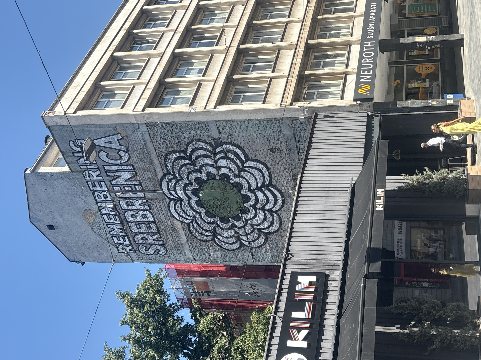 In Sarajevo, Bosnia and Herzegovina, a mural on the side of a building reads "REMEMBERING SREBRENICA" above a stylized floral design. To the left, a tree partially obscures scaffolding on another building covered in red netting. Below the mural, a storefront displays the "KLIM" logo. Further to the right, another building houses businesses including "NEUROTH SLUŠNI APARATI". Two individuals are walking on the sidewalk in front of the storefronts; one is a person in a yellow dress, and the other is a person in a white shirt and dark pants. The sky is clear and blue.