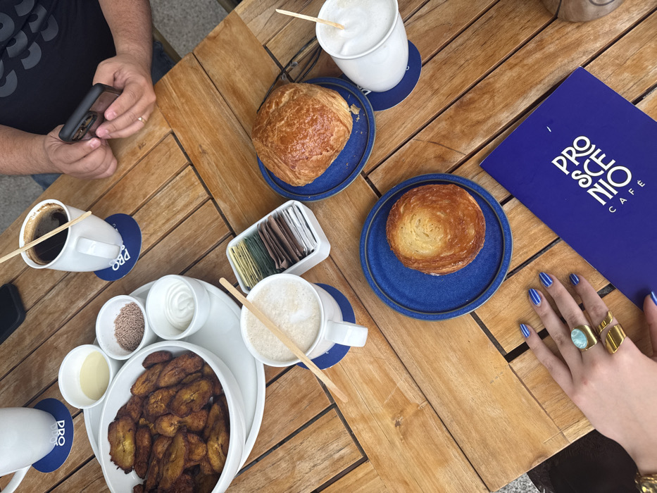 An overhead view depicts a wooden slatted table at Proscenio Café in Mexico City, Mexico. The table is laden with breakfast or snack items for at least two individuals.
**Objects:**
* A large white oval bowl contains numerous fried plantain slices, resting on a larger white serving tray.
* Three small white bowls accompany the plantains, holding a light yellow liquid (condensed milk), brown sprinkles, and a white cream.
* Four white mugs are present: one partially visible at the top left, one containing black coffee with a wooden stirrer at the top right, and two containing foamy, latte-style beverages with wooden stirrers in the mid-left and mid-right. All visible mugs sit on round blue coasters branded with "PRO".
* A white rectangular caddy holds various individual sugar and sweetener packets.
* Two round blue ceramic plates display two golden-brown pastries of different sizes, possibly croissants or similar laminated dough items.
* A pair of dark-rimmed eyeglasses rests near one pastry.
* A blue menu or booklet with "PROSCENIO CAFÉ" printed in white text is visible in the lower right quadrant.
**People:**
* A person's hand (identified as male by a ring on the left ring finger) holds a black-cased smartphone in the upper right.
* A second person's hand (identified as female by blue striped nail polish and two gold rings, one featuring a large teal-colored stone) rests on the table in the lower left.
**Environment:**
The scene is set on an outdoor wooden table, indicated by the slatted pattern and direct lighting. The immediate background is out of focus, suggesting an urban patio or sidewalk cafe environment.