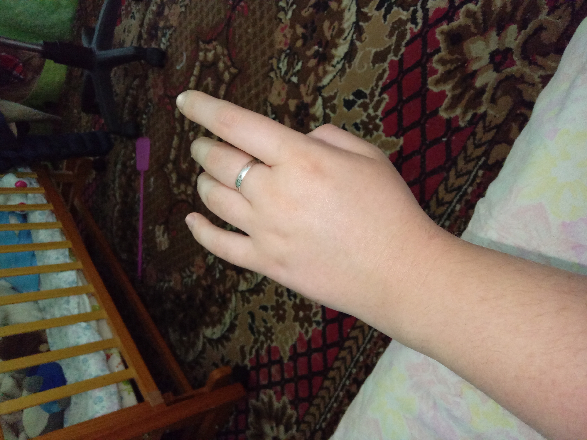 The image shows a close-up view of a person's arm and hand, resting on a patterned carpet. The person is wearing a silver ring on their left ring finger. In the background, a wooden crib with bedding and toys can be seen. A black office chair and a pink object on the floor are also visible. The scene appears to be indoors, with the lighting suggesting it is daytime. The location is identified as Kitskany, Moldova.