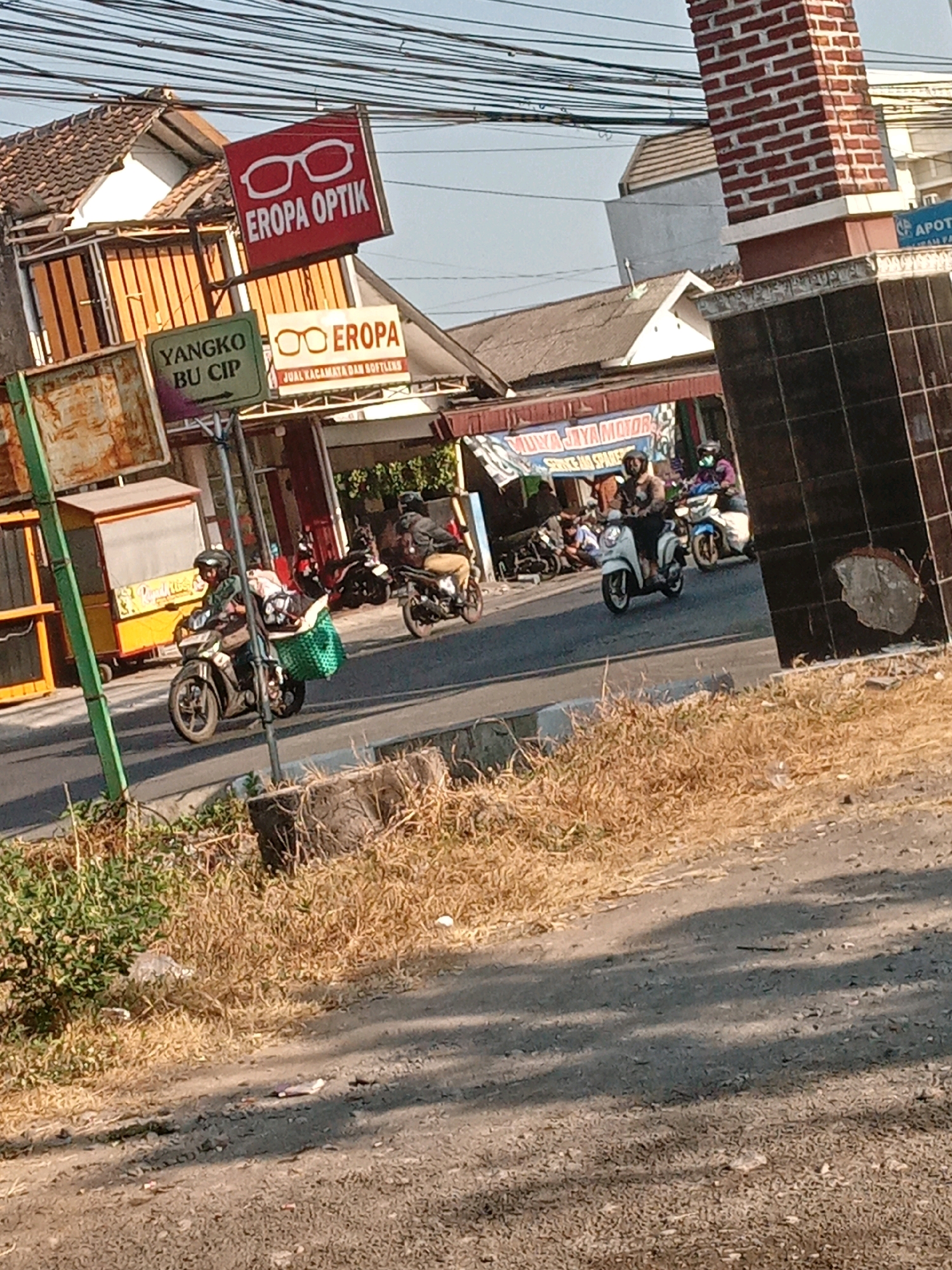 In Banguntapan, Indonesia, a street scene unfolds under a bright sky. Several motorcycles are visible, with four riders and their vehicles prominent. One motorcyclist in the foreground wears a helmet and a brown jacket, with a large green woven basket secured to the rear of their scooter, carrying luggage. Another rider behind them wears a helmet and a brown jacket, with a passenger also wearing a helmet. To the right, a rider in a purple shirt and helmet is on a white scooter, and further back, another rider in a black jacket is on a dark motorcycle.
Signs are present in the scene, including a red sign with white text reading "EROPA OPTIK" and an image of eyeglasses, and a green sign with white text reading "YANGKO BU CIP". Another sign displays "EROPA" with eyeglasses and "JUAL KACAMATA DAN SOFTLENS". A banner with "MUYA JAYA MOTOR" and Indonesian text is visible behind the motorcycles.
The environment includes a paved road, dry grass and vegetation along the roadside, and a brick structure with dark tiled accents on the right. Buildings with tiled roofs are visible in the background, along with overhead power lines. The overall scene depicts a typical street in a commercial or residential area of Banguntapan.