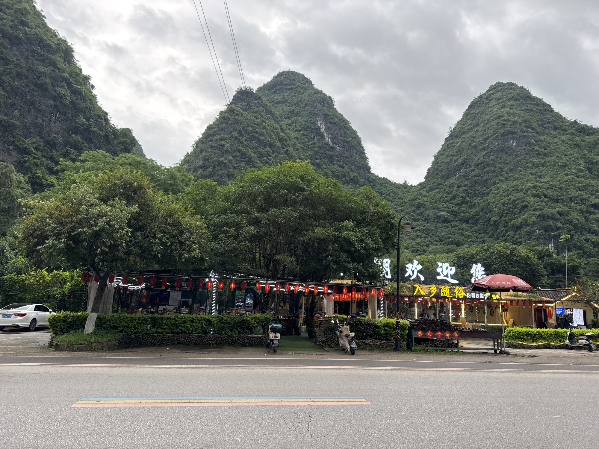 The image depicts a scene along a road in Guilin, China, with distinctive karst mountains in the background under an overcast sky. In the foreground, a road with colored stripes stretches across the frame. Adjacent to the road is a building or establishment, possibly a restaurant, adorned with red lanterns and illuminated Chinese signage. Scooters are parked in front of the building. A white car is parked to the left. The building is surrounded by greenery, including trees and bushes. Power lines are visible above the mountains.