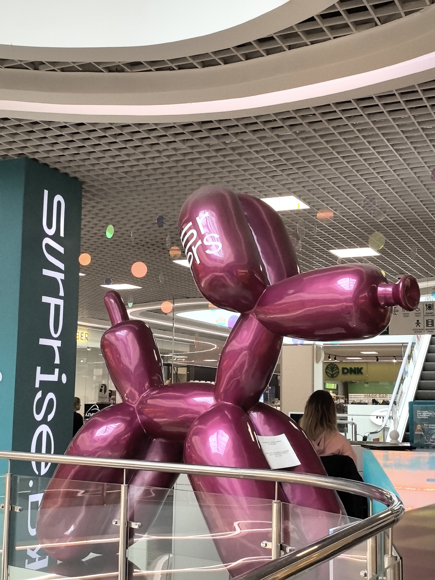 In the city of Unknown, Unknown, a large, shiny, metallic pink balloon dog sculpture is displayed indoors, likely in a shopping mall. The sculpture is positioned in the foreground, taking up a significant portion of the frame. It has a glossy finish that reflects the surrounding environment, including lights and architectural elements.
The background reveals a multi-level indoor space with a patterned ceiling made of square panels. Fluorescent lights are visible in the ceiling, casting a bright illumination. Several colorful, translucent circles of varying sizes are suspended from the ceiling, adding a decorative element to the space.
To the left of the sculpture, a tall teal-colored pillar features the white text "surprise" written vertically. In the mid-ground, a woman with long, light brown hair is seated, facing away from the camera, wearing a light pink hooded sweatshirt and dark bottoms. She is positioned near a railing with chrome accents and glass panels.
Further in the background, there are escalators and shop signage, including a green sign with a logo and the text "DNK". Other store signs and displays are also visible. The overall environment suggests a modern, commercial setting, likely a shopping center, where the art installation is a focal point.