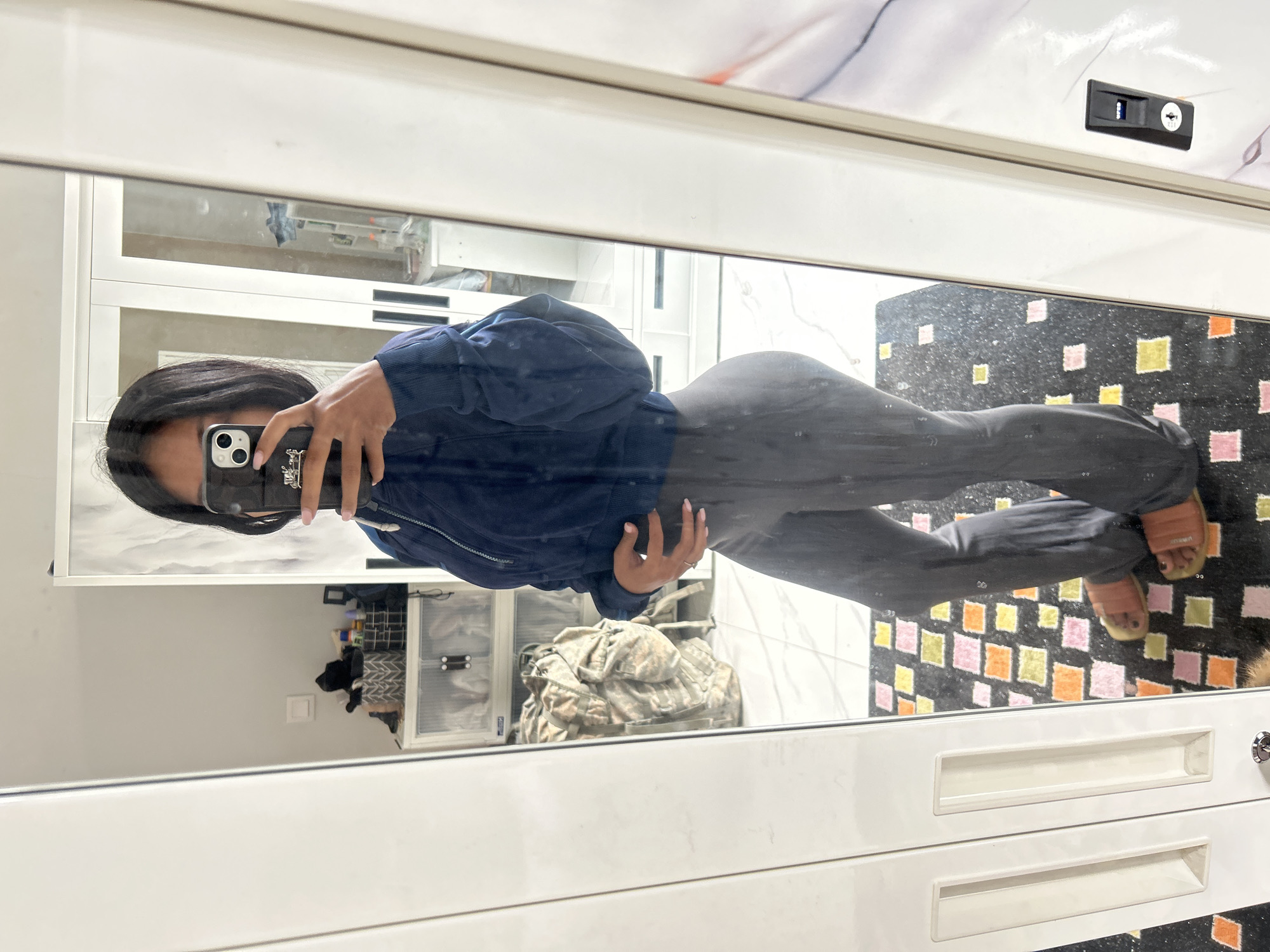 A person is taking a mirror selfie in what appears to be a room with white cabinets and a marble-patterned ceiling. The person is wearing a dark blue jacket and dark pants, and is holding a phone in their right hand, which is angled to capture their reflection. The phone case has a detailed design on it. The reflection shows the person's face and hair, along with their legs and feet, which are clad in orange slides. In the background, reflected in the mirror, are various objects including storage bins, a camouflage-patterned bag, and a rug with a colorful, checkered design. A black electrical outlet with a keyhole is visible on the upper right side of the mirror's frame. The lighting suggests it is daytime, and there are no obvious clues about the weather or specific emotion being conveyed. The location context is Sukawati, Indonesia.