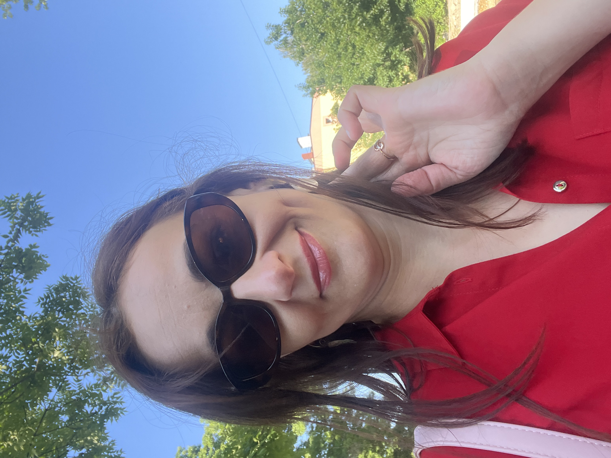 A woman is taking a selfie outdoors, with a bright blue sky and green trees in the background. She is wearing a red top and sunglasses. Her right hand is raised to her head, and she is smiling. A silver ring is visible on her finger. The lighting suggests it is daytime, likely sunny. The background appears to be a residential or park area, with a building and trees visible. The image is taken from a low angle, looking up towards the woman and the sky.