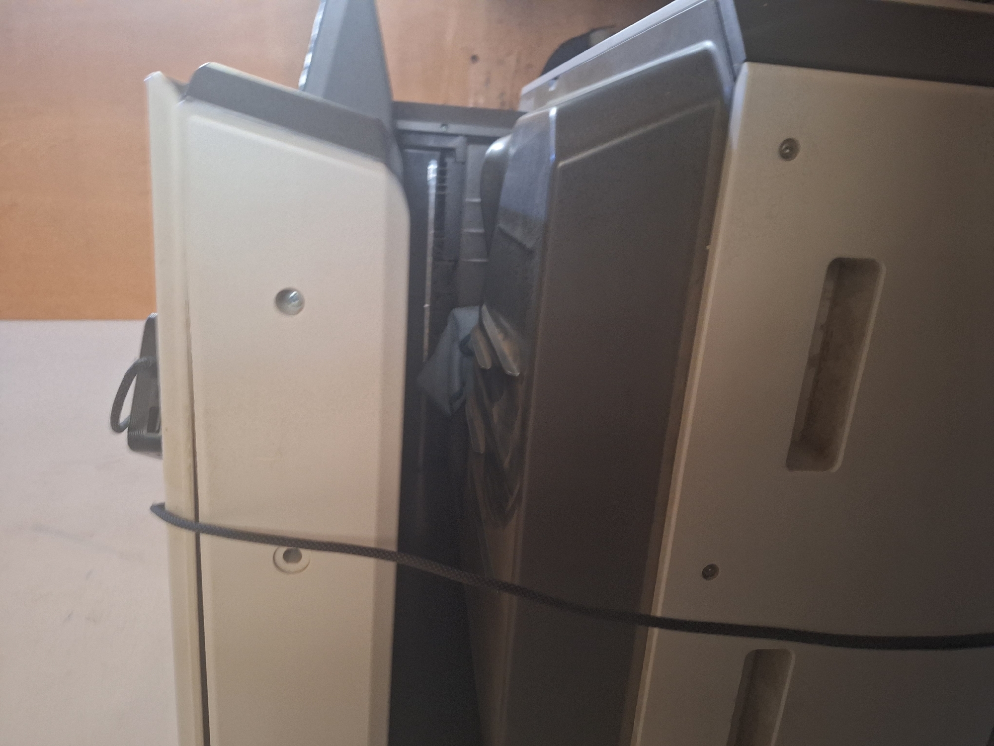 The image shows a close-up, angled view of a large, off-white and dark gray office machine, possibly a printer or copier, situated indoors. A black power cord dangles from the top of the machine. The machine appears to be dusty, with visible smudges and a thin layer of dust on its surfaces. The background consists of an out-of-focus wall painted in neutral tones. The lighting is dim and appears to be artificial. No people are visible in the frame. The setting does not provide specific clues to identify the exact location within Jalingo, Nigeria.
