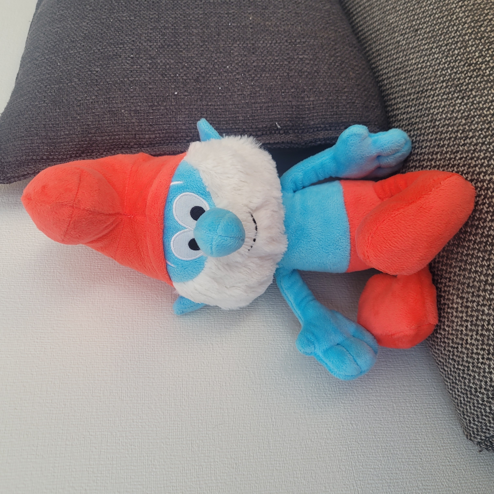 A plush toy of Papa Smurf is sitting on a couch next to a wall. The toy is blue with a white beard, a red hat, and red pants. The couch is dark grey and the wall is white. The scene is indoors, and the lighting is natural. The image is taken from a slightly high angle, looking down at the toy. The background is slightly blurred, drawing attention to the toy. The image appears to have been taken in a residential setting, possibly a living room. There are no people visible in the image. There is no information in the image to suggest it is located in Jūrmala, Latvia.