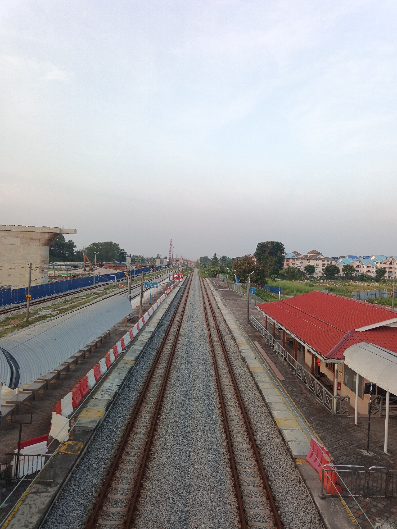 An elevated view captures a train station in Klang City, Malaysia, under a pale, overcast sky. The scene is dominated by a pair of parallel train tracks stretching into the distance, flanked by gray gravel. To the left, a covered platform features a corrugated metal roof and benches, with red and white safety barriers along its edge. Construction is evident on the far left, with concrete structures and blue fencing. To the right, a red-roofed building, likely a station house, has a covered walkway and stairs. Beyond the station, residential buildings with blue and cream facades are visible amidst greenery. The overall impression is one of ongoing development and a typical urban environment. There are no people visible in the image.