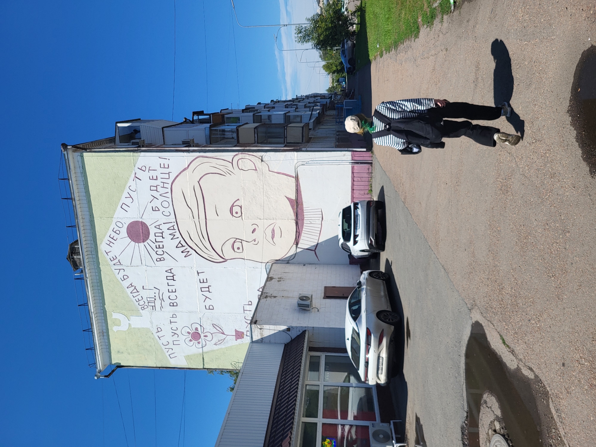 On a sunny day, a young person with green-dyed hair, wearing a striped shirt, black pants, and a backpack, walks away from the camera down a sidewalk in the city of Petropavl, Kazakhstan. The sidewalk is paved and appears to be bordered by grass on the right. To the left, a large apartment building dominates the scene, adorned with a mural. The mural depicts a child's face and includes text in Russian, along with drawings of a sun, a flower, and a bird. The building has balconies on its side and appears to be in a residential area. In front of the building, two white cars are parked. One is a sedan, and the other is an SUV. The sidewalk and road show some wear and tear, with a puddle of water visible on the pavement. In the background, more apartment buildings and trees can be seen, and the sky is a clear, bright blue.