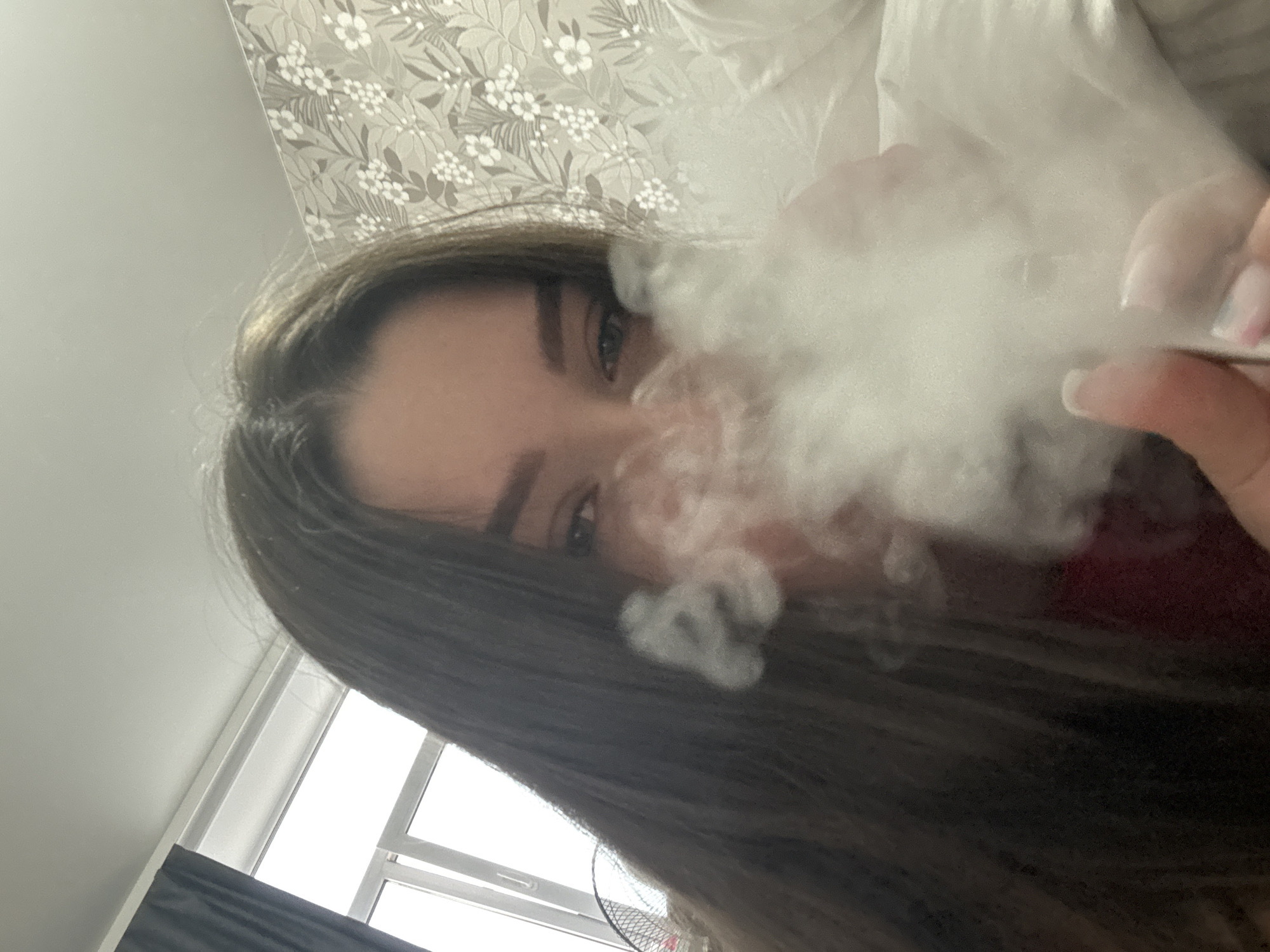 An indoor, low-angle selfie captures a young woman exhaling a large cloud of white smoke or vapor, likely from a vaping device partially visible in her right hand. Her long dark brown hair frames her face, with her eyes and eyebrows visible above the dense smoke. She appears to be looking directly at the camera. Her right hand shows manicured fingernails with light-colored polish.
The background suggests a domestic setting. In the upper portion of the frame, there is patterned wallpaper with a grey or beige floral design featuring white flowers and leaves. Below this, behind the smoke, white bedding or pillows are visible. To the lower left, a window with white frames and a dark curtain allows natural light into the room, suggesting daytime. A portion of a black fan is also visible in the lower central background. The overall impression is a casual, personal moment indoors, likely in a bedroom. No text is visible in the image.