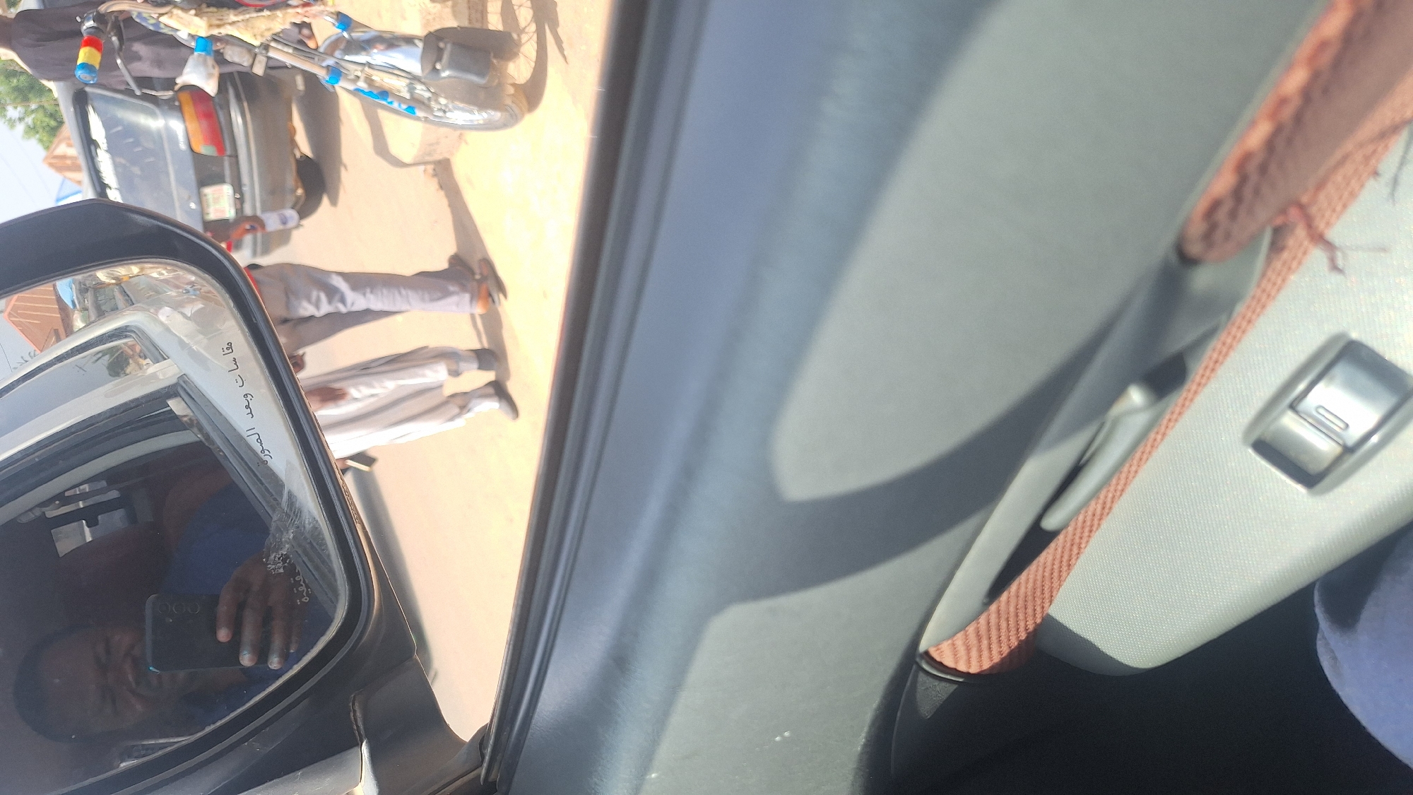 In the city of Jalingo, Nigeria, a man is captured in a reflection of a car's side mirror, holding a smartphone and taking a selfie. He is wearing a blue shirt. Through the mirror, other individuals are visible: one person wearing a white traditional outfit, another in grey trousers and a darker shirt, and a third partially obscured. In the background, a black sedan is parked, and a motorcycle is also present. The setting appears to be an outdoor, urban environment, possibly a street or roadside, with a dusty ground. The interior of the car, including a door panel with a fabric insert and a window control button, is also visible in the foreground.