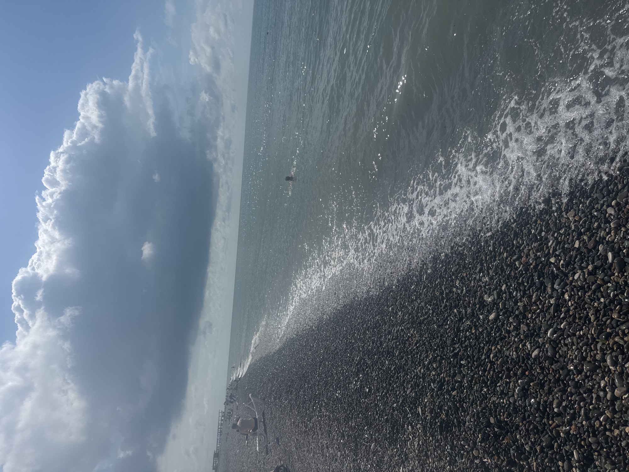 This image depicts a beach scene in Batumi, Georgia. The foreground is dominated by a dark pebble beach, wet from the lapping waves. The sea stretches out to the horizon, its surface shimmering with sunlight. A few people are visible on the beach, some seated on chairs and others walking. In the water, a single person can be seen swimming or wading. The sky is partly cloudy, with large, dramatic clouds casting shadows on the sea and beach. The overall atmosphere is one of a relaxed summer day at the coast. The time of day appears to be late morning or early afternoon, given the brightness of the sun and the shadows. There is no visible text in the image.