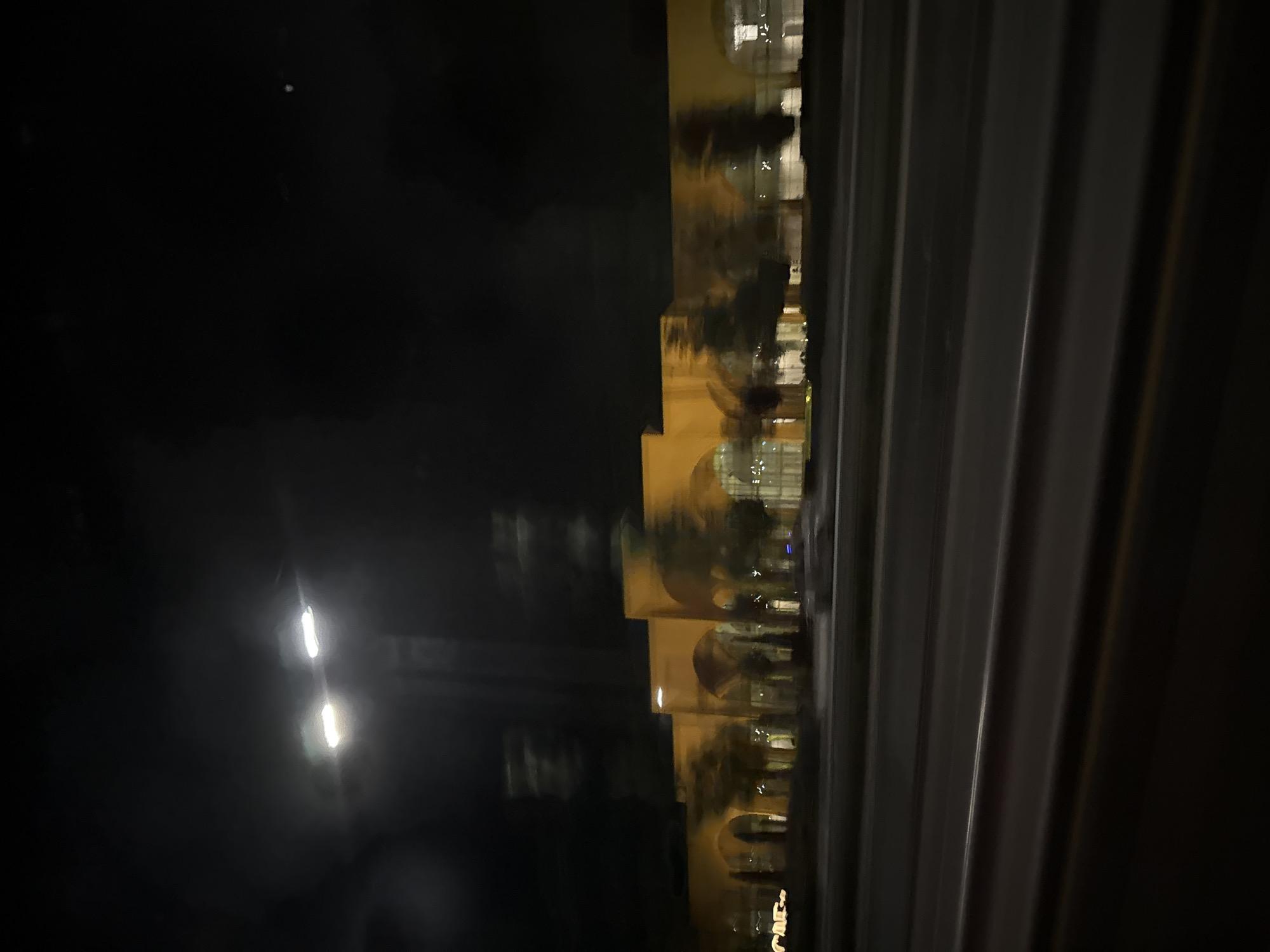 A night-time, blurred photograph captures a long, low-rise building with repeating arched windows, illuminated by interior lights. The building's facade is a warm yellow hue. Trees are visible in front of the building, appearing as dark, indistinct shapes. In the foreground, blurred horizontal lines suggest a road or highway, indicating the photo was taken from a moving vehicle. Two bright streetlights are visible in the upper left of the image, their light also appearing blurred. The overall environment is dark and urban. The building is located in Tukaytepa, Uzbekistan.