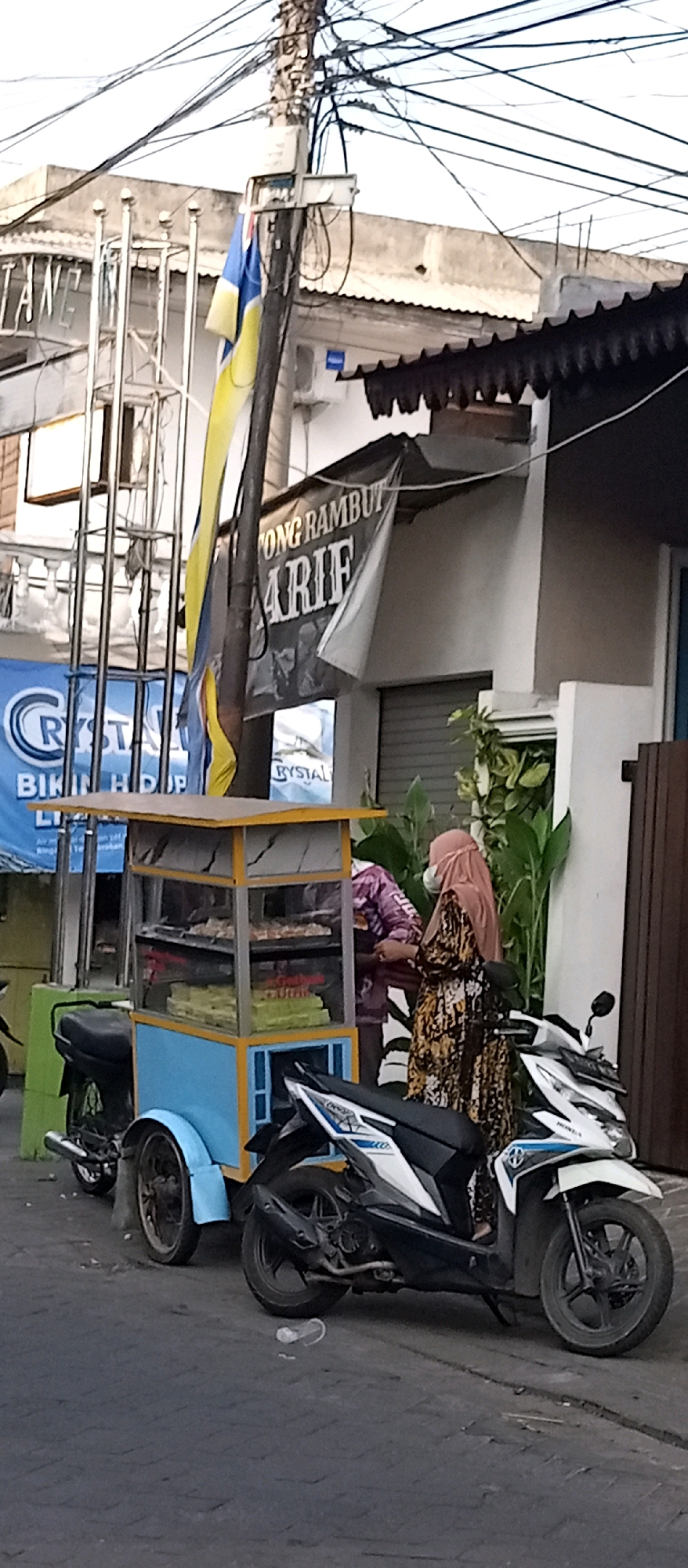 The image depicts an outdoor street scene in Wadungasri, Indonesia, featuring a food cart parked beside two motorcycles. A woman wearing a headscarf and a patterned dress stands at the food cart, seemingly interacting with another person who is partially obscured. The food cart itself is blue and yellow and contains several shelves of food items, suggesting it is a mobile food vendor. Behind the cart and people, a building with a dark-tiled roof and a banner that reads "TONG RAMBUT ARIF" is visible. The environment is urban, with a paved street and overhead electrical wires, indicating a typical street setting within a populated area.