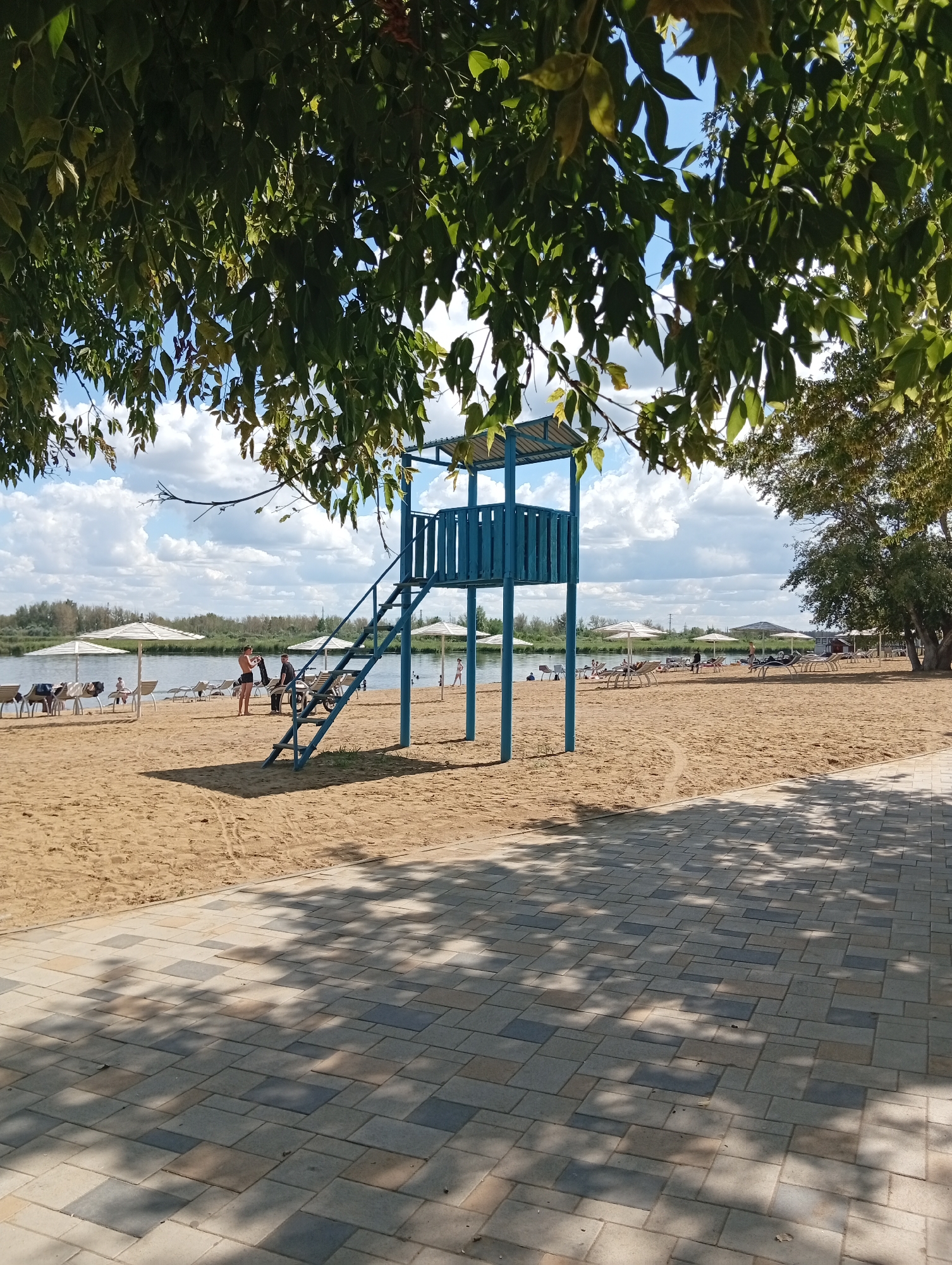 The image depicts a beach scene under a cloudy sky. A blue lifeguard tower with stairs is prominently featured in the foreground, positioned on a sandy beach. Several white beach umbrellas and lounge chairs are scattered across the sand. The water is visible in the background, with trees lining the far bank. A paved walkway with a stone pattern runs along the bottom right of the image, casting dappled shadows from overhead foliage. A few people are visible on the beach, some standing near the lifeguard tower, others near the water or resting on chairs. The overall impression is one of a recreational area by a body of water. The location is identified as Мичурин ауылдық округі, Kazakhstan.