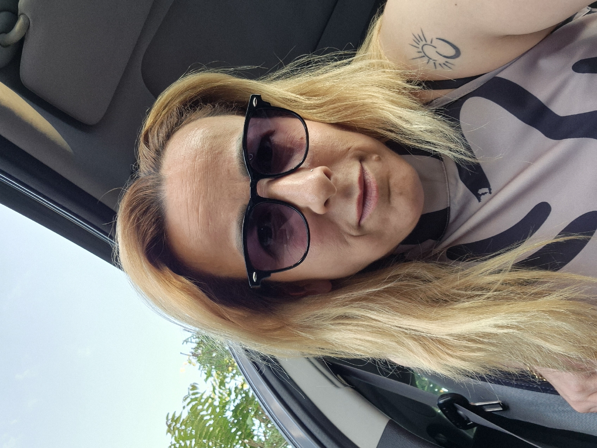 The image displays an adult individual, appearing female, with light skin and shoulder-length blonde hair, seated inside a vehicle in the city of Unknown, Unknown. The person is wearing dark-rimmed sunglasses with dark tinted lenses. A small silver stud is present in the left nostril. Visible lines are on the forehead and around the eyes, and a small mole is on the chin. The individual is wearing a sleeveless top or dress with a light beige base color and large, irregular dark brown or black abstract patterns. A black tattoo on the upper left arm depicts a stylized sun-like symbol positioned above an interlocking or crescent-shaped design. The person is looking directly towards the camera. The vehicle interior, visible on the left and above the person, is dark grey or black, with a seatbelt visible to the left. Through the open roof or window, a bright, clear light blue sky with some wispy clouds and green foliage from trees or bushes are visible, indicating an outdoor, daytime environment.