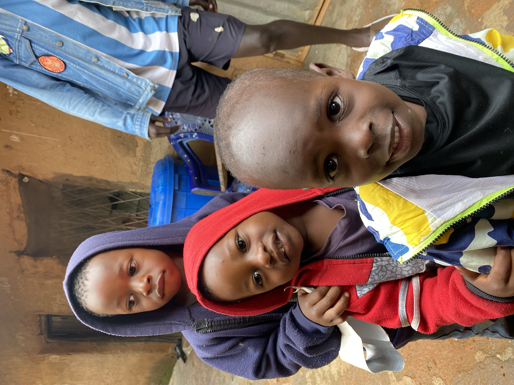 Four children are shown in a medium shot, standing outside a building with rough-textured, tan-colored walls and a barred window. The child in the foreground on the right is looking directly at the camera, with a light-colored, patterned jacket over a black t-shirt. To their left, a child in a red hooded jacket is looking towards the left of the frame. Behind them, a child in a dark blue hooded jacket is also looking to the left, their hood obscuring part of their face. In the background, an adult is partially visible, wearing a denim jacket over a blue and white striped shirt and dark shorts. A blue plastic container and a chair are also present in the background. The ground appears to be uneven and muddy. The overall setting suggests an outdoor area in a residential or communal space, likely in Jos, Nigeria, given the context.