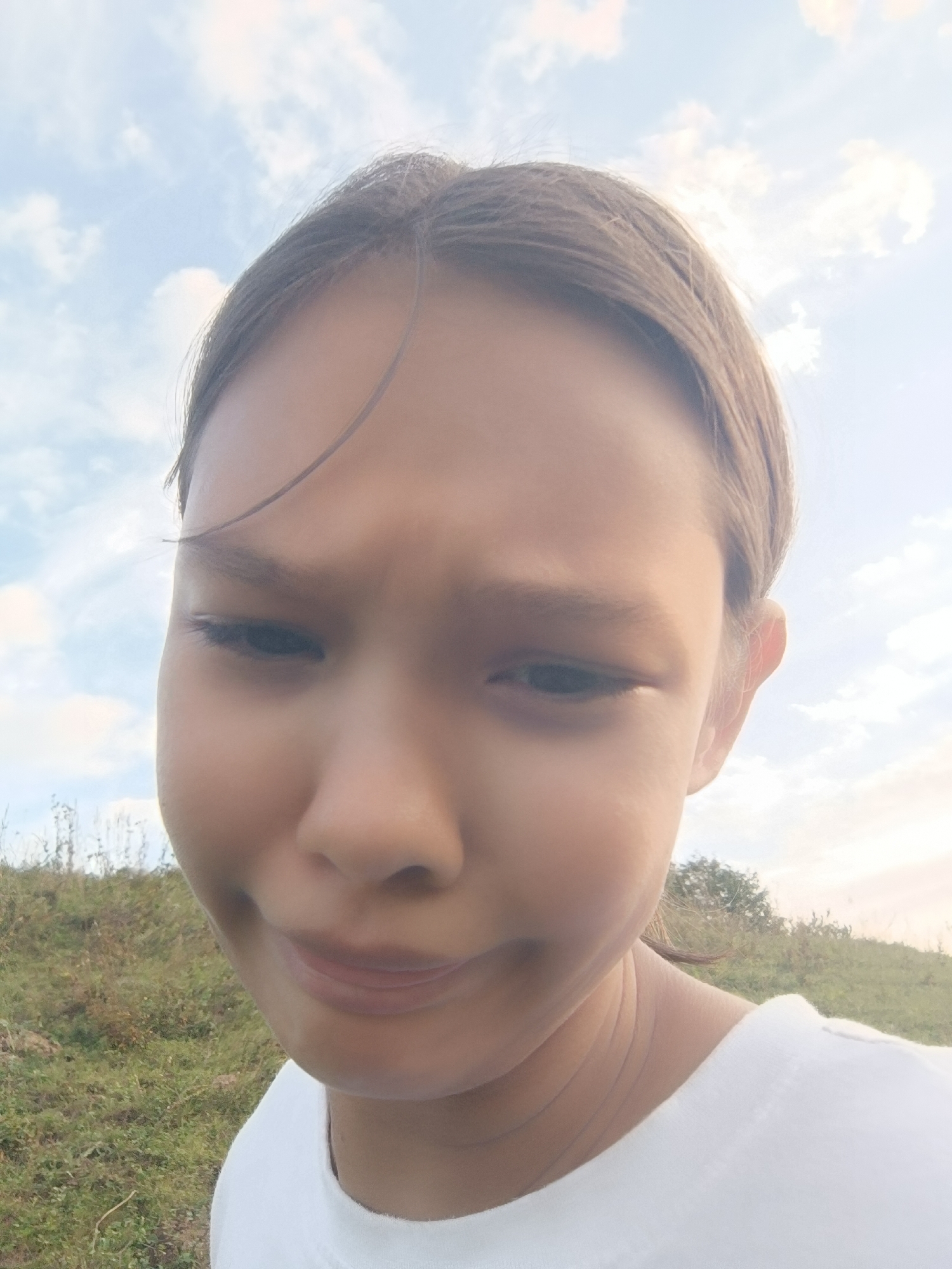 The image shows a close-up, waist-up, outdoor selfie of a young person with light brown hair, looking down and slightly to the right. The person is wearing a white t-shirt. The background consists of a grassy, hilly landscape under a bright, cloudy sky. The specific location within the city of Karsha, Russia is not discernible from the visual information provided.