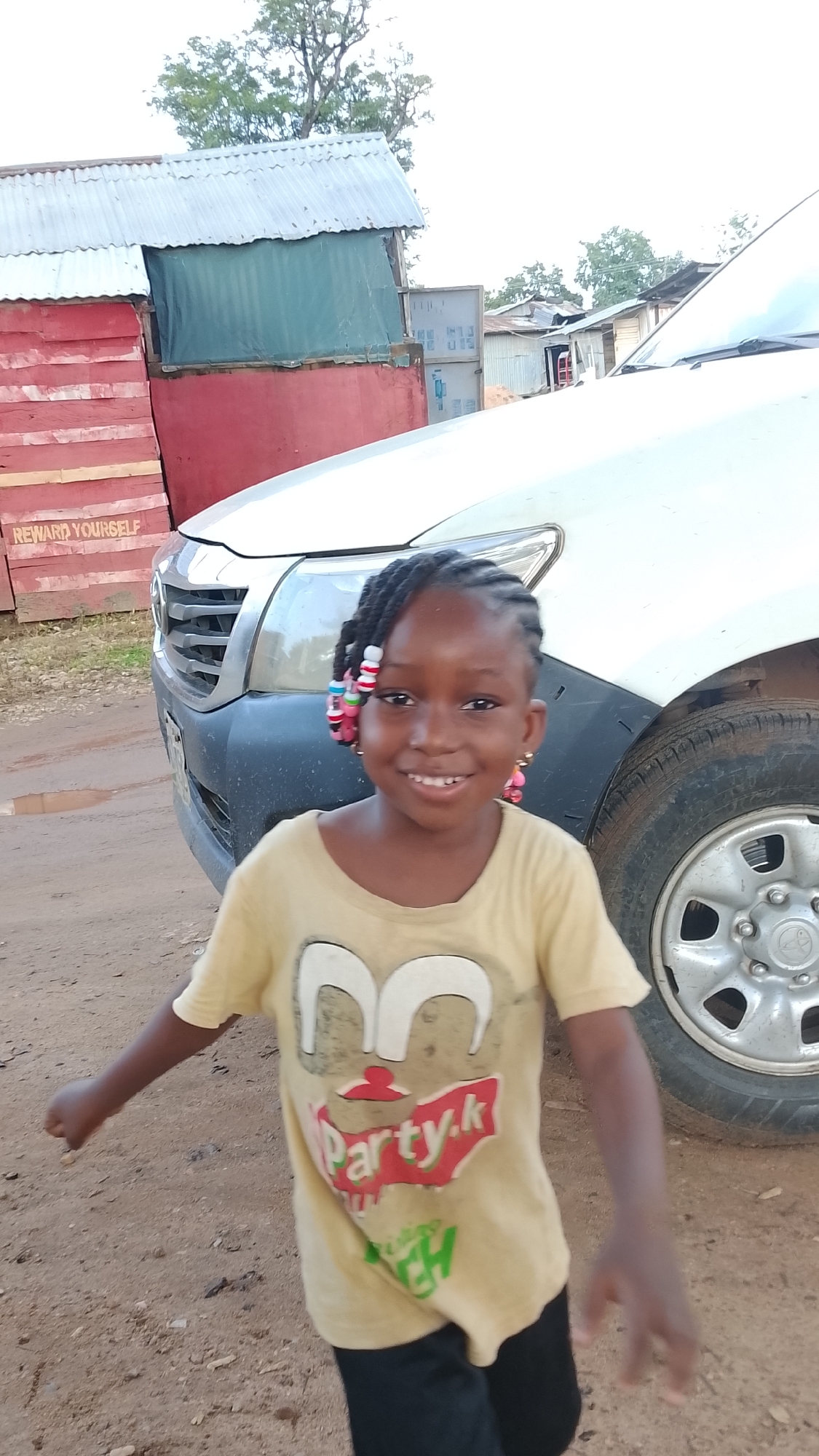 A young Black girl with braided hair adorned with colorful beads and pink earrings smiles at the camera. She is wearing a light yellow t-shirt with a graphic print of a face and the words "Party.k" and "CH" in green. The girl is standing in front of a white vehicle, with its grille and headlight visible. Behind the vehicle, there are structures made of corrugated metal and red wooden planks. Text on the red planks reads "REWARD YOURSELF". The ground is dirt. The scene appears to be an outdoor setting in Adankolo, Nigeria.