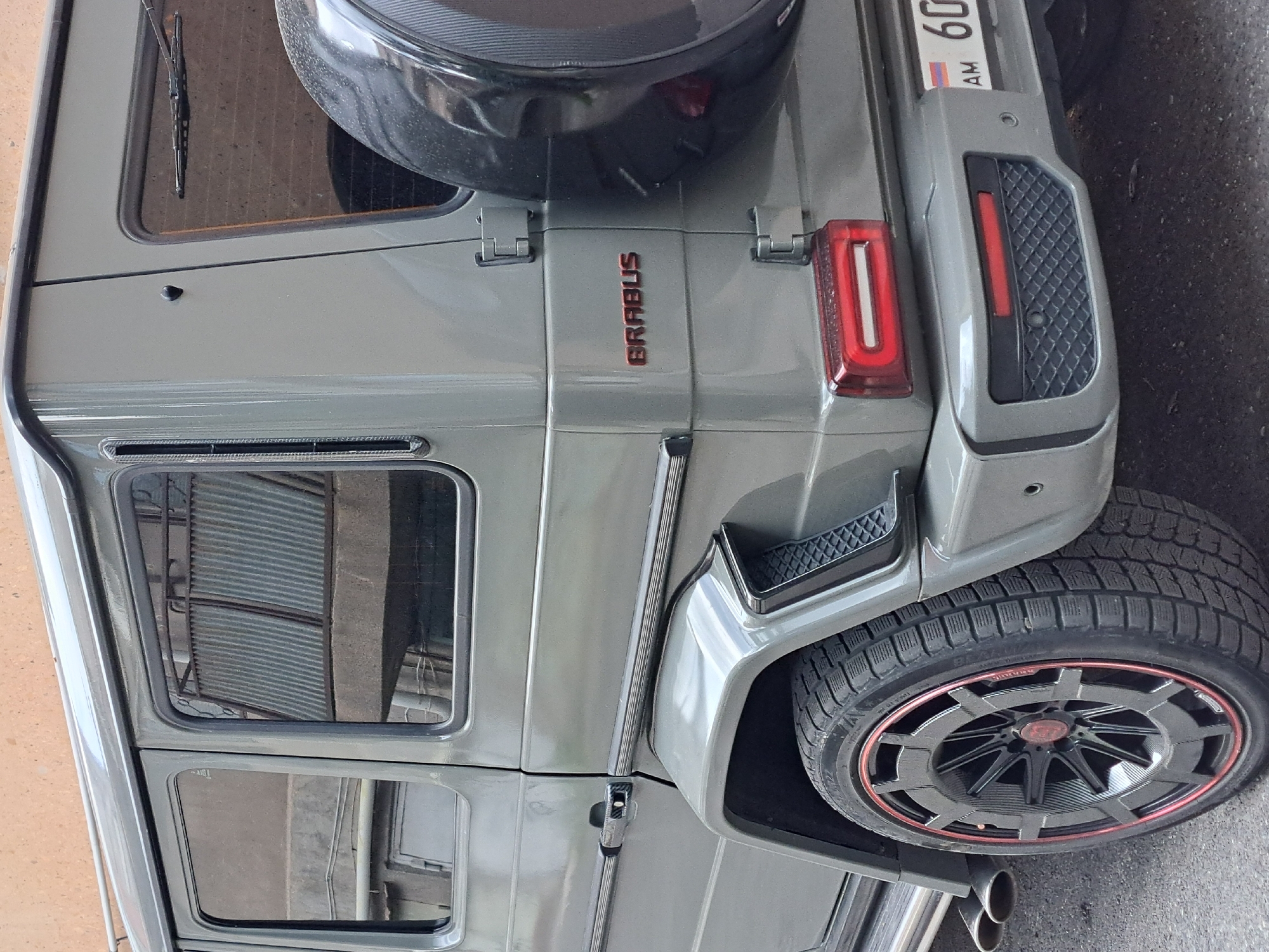 A gray Brabus G-Wagon is parked in Yerevan, Armenia. The vehicle has a spare tire mounted on the rear door. The license plate reads "60 AM." The wheels are custom black and red. The location appears to be a street.