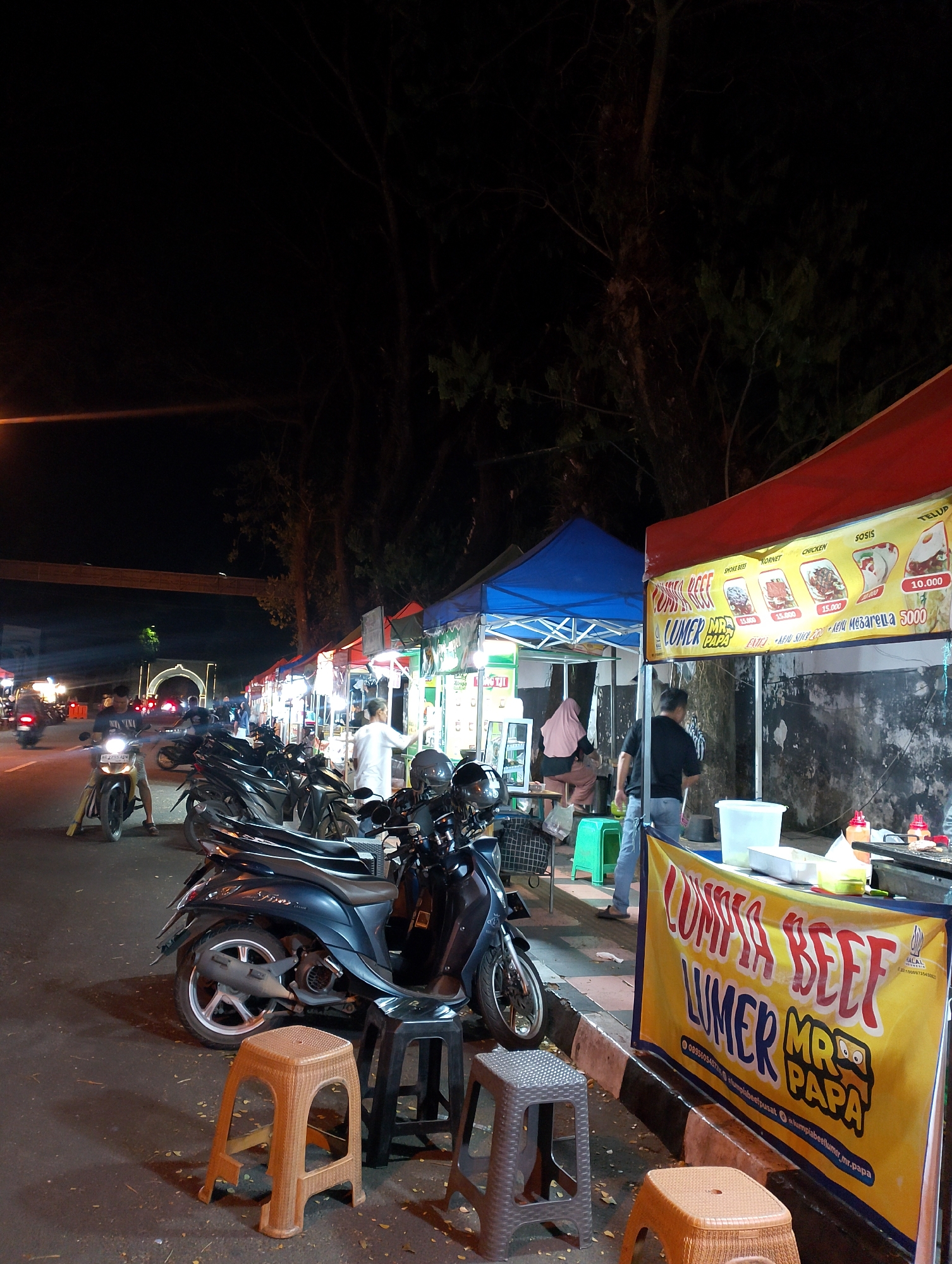 A street-level, night-time, vertical photograph captures a vibrant street food scene in Magelang, Indonesia. The scene is dominated by a row of food stalls lining a street, characterized by their brightly colored awnings. A red awning stretches across the top right of the image, displaying a menu board with prices. To the left, a blue awning is visible. Parked motorcycles, a common mode of transport in the region, fill the foreground and spill onto the road. Several people are present, including a man in a white shirt serving customers, a man in a black shirt and jeans, and two women dressed in a pink headscarf and a grey headscarf respectively, seated at tables. The street is paved, and the background features dark trees and a pedestrian overpass illuminated by lights, suggesting a busy urban environment. The overall atmosphere is lively, with the glow of stall lights and the hum of nighttime activity creating a warm and inviting ambiance.